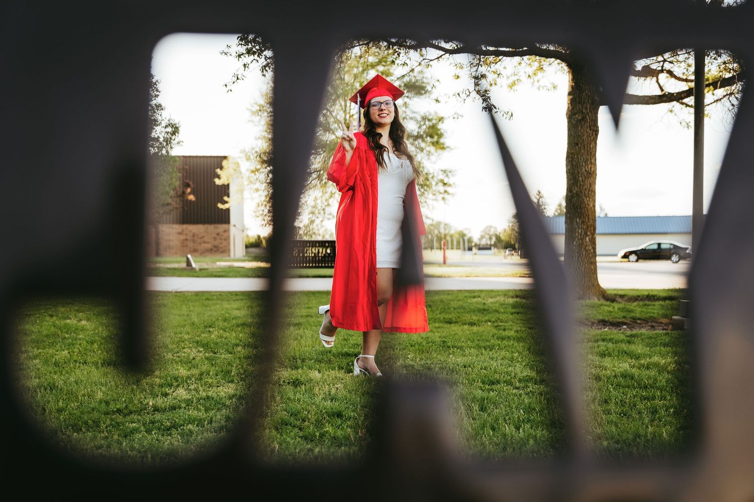Graduate in red cap and gown poses for photos on a sunny day near a lake.