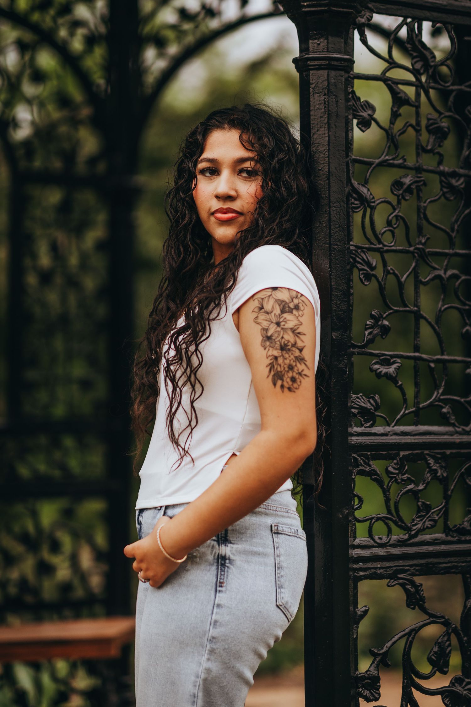 A person wearing a white top and light denim overalls poses outdoors against an ornate iron gate with greenery.