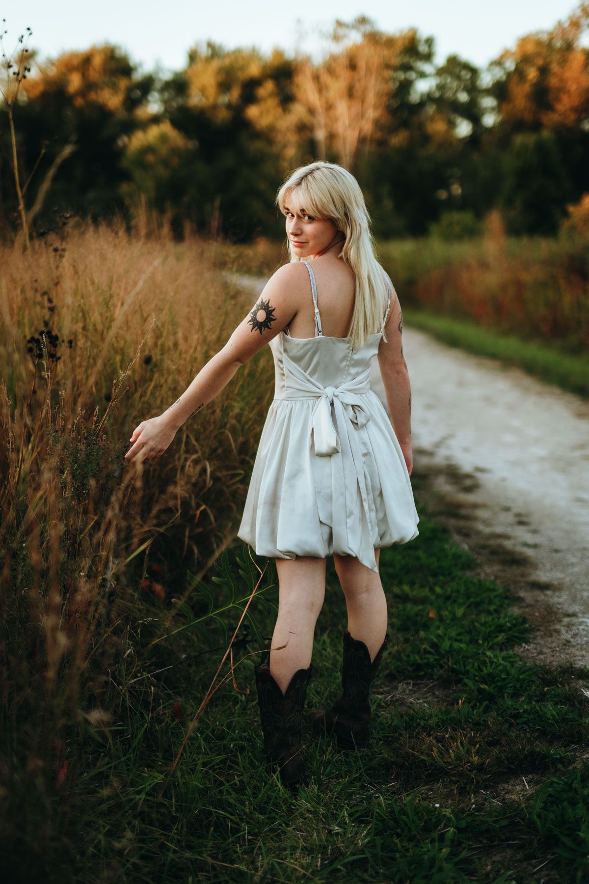 A person in a white sundress and cowboy boots walks along a dirt path during golden hour with autumn foliage in background.