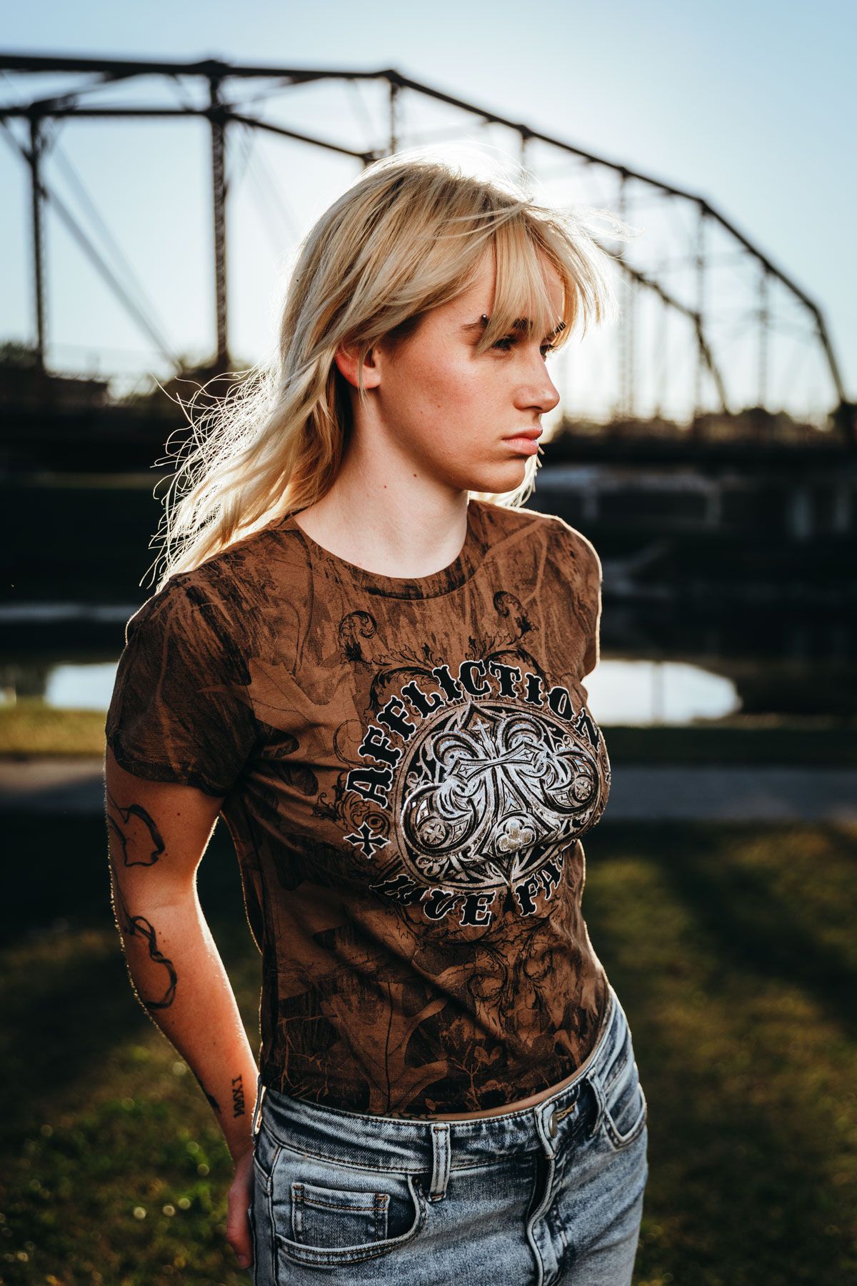 Person in a brown graphic t-shirt with circular design poses near metal bridge structure at sunset.