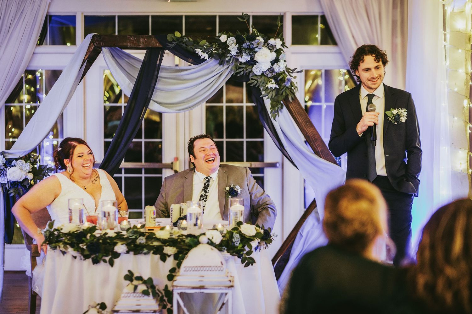 A wedding speech being delivered at an elegantly decorated indoor venue with draped fabric and floral accents.