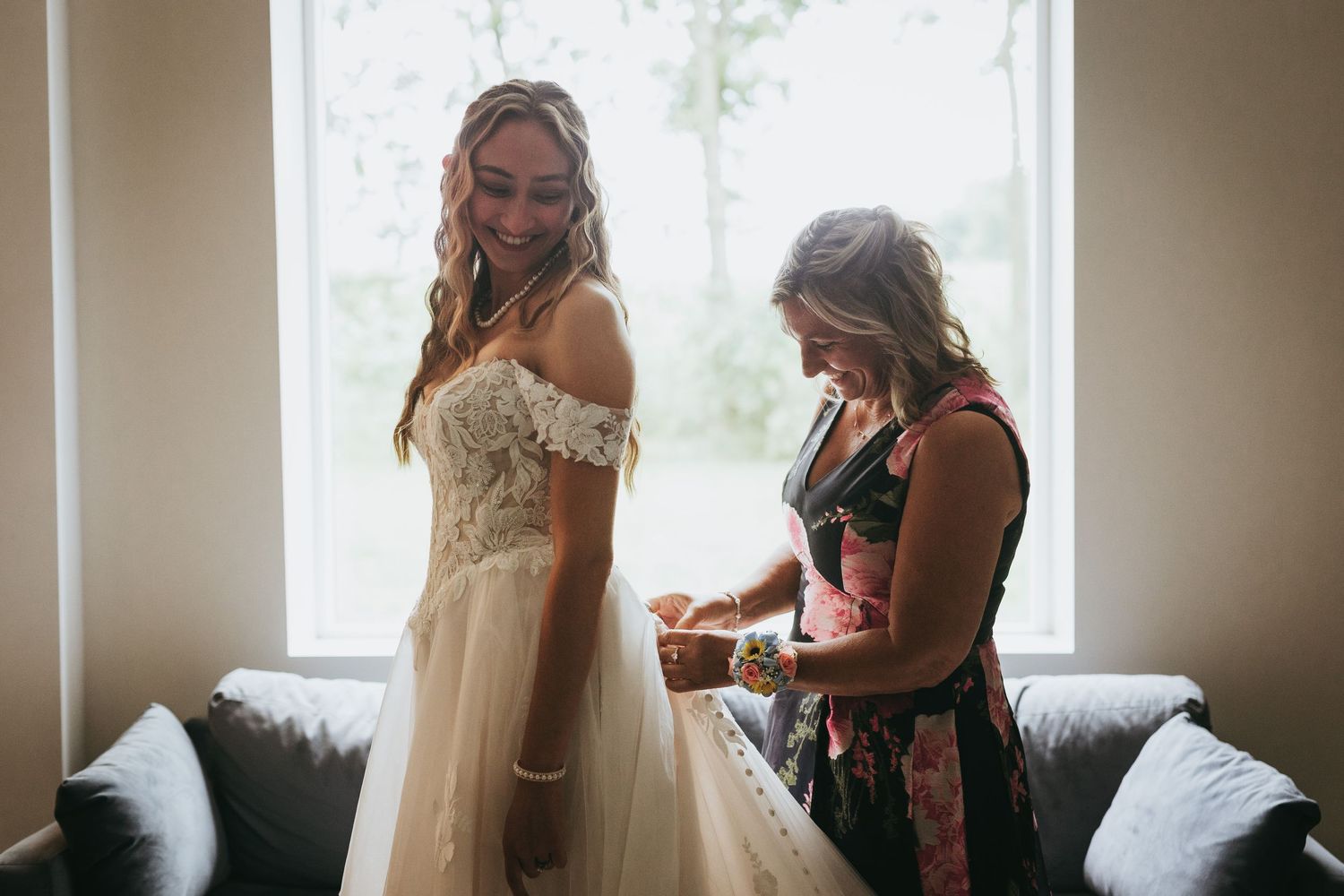 Bride smiling in lace off-shoulder wedding gown while woman helps fasten dress near bright window.