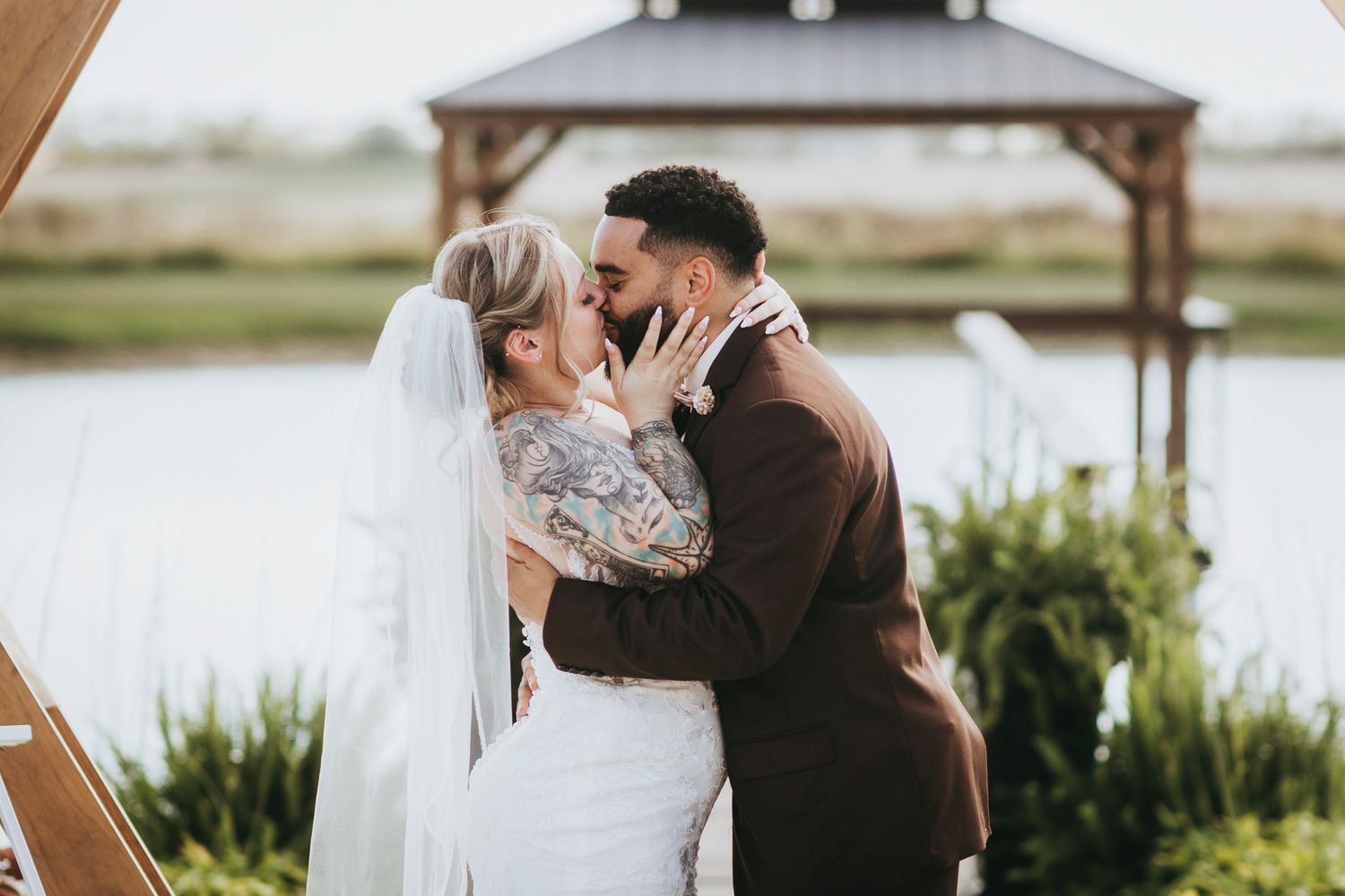 A couple shares a romantic moment during their wedding photoshoot by a scenic lake with greenery in the background.