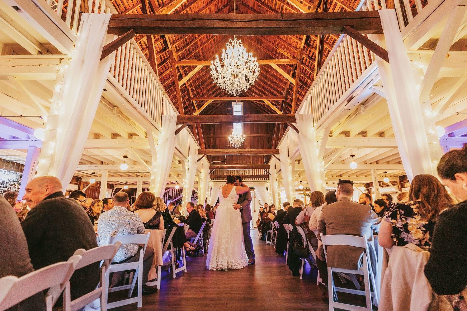 A wedding ceremony in a grand barn venue with high wooden beams and chandeliers.