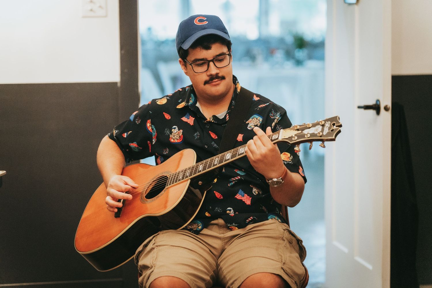 Man in blue cap and floral shirt playing acoustic guitar while seated indoors.