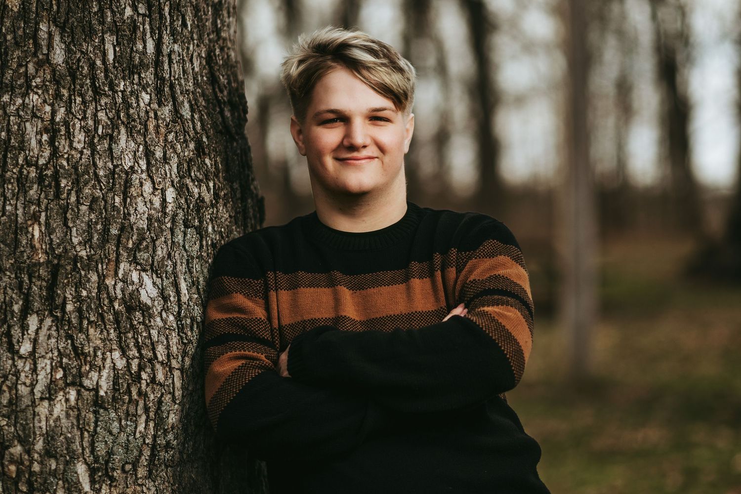 Young man in striped sweater leaning against a tree with arms crossed, smiling outdoors in a wooded setting.