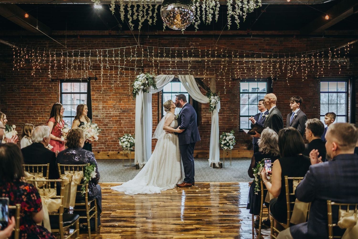 Indoor wedding ceremony in a rustic brick venue with string lights and white floral arch decorations.