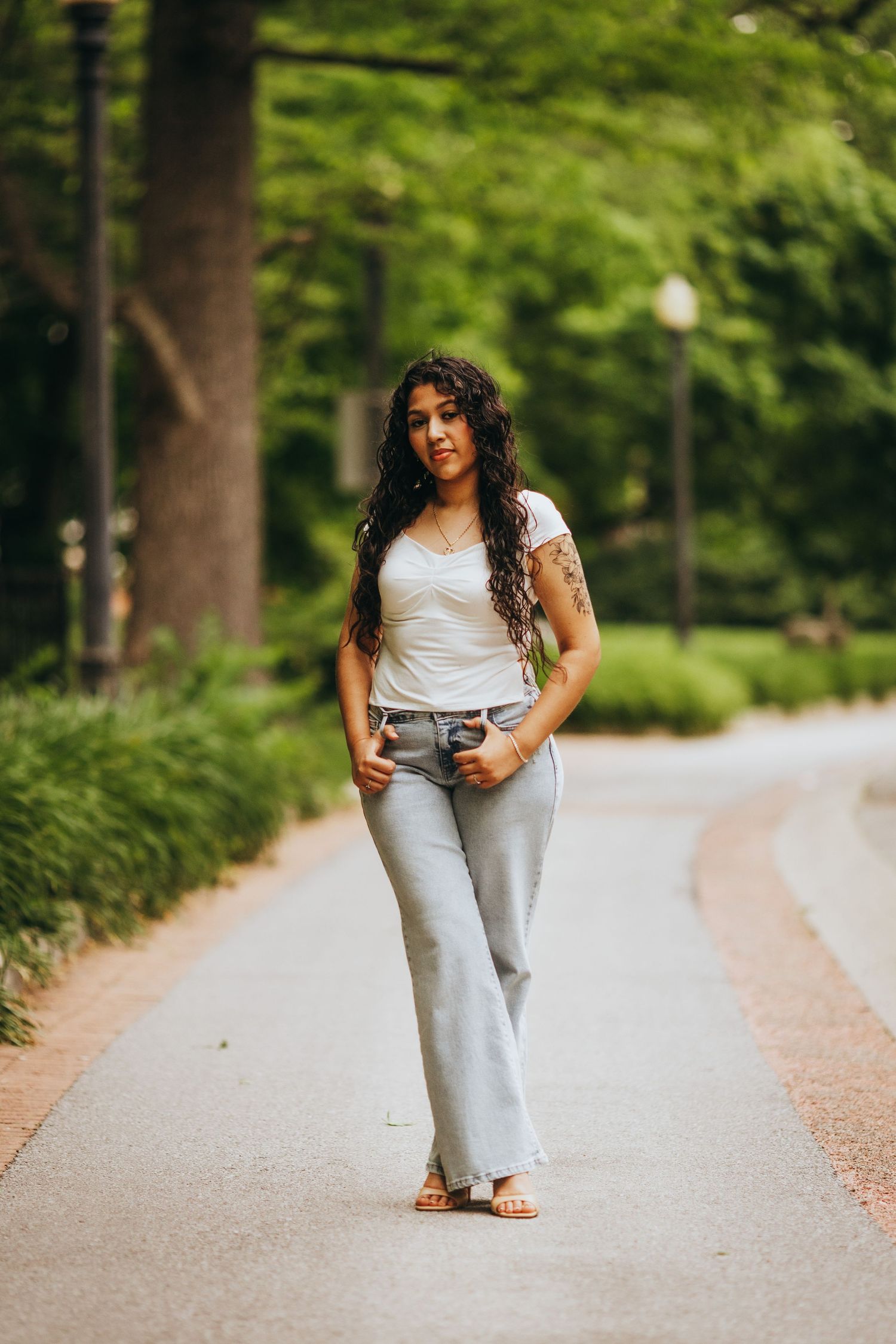 A person in a white top and light jeans poses casually on a tree-lined path in a park setting.