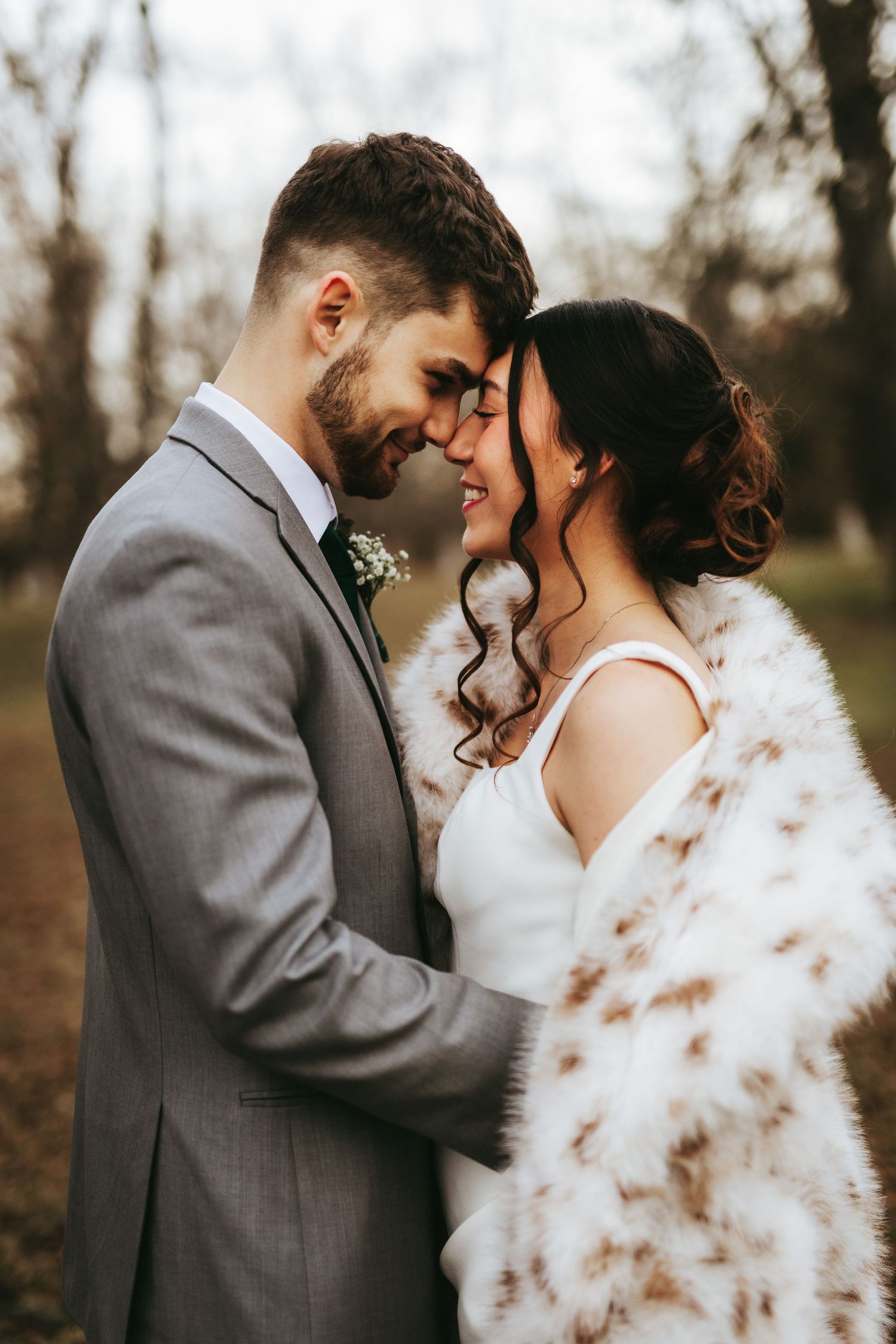 Romantic wedding couple in autumn setting with bride wearing white fur wrap and groom in gray suit sharing intimate moments.