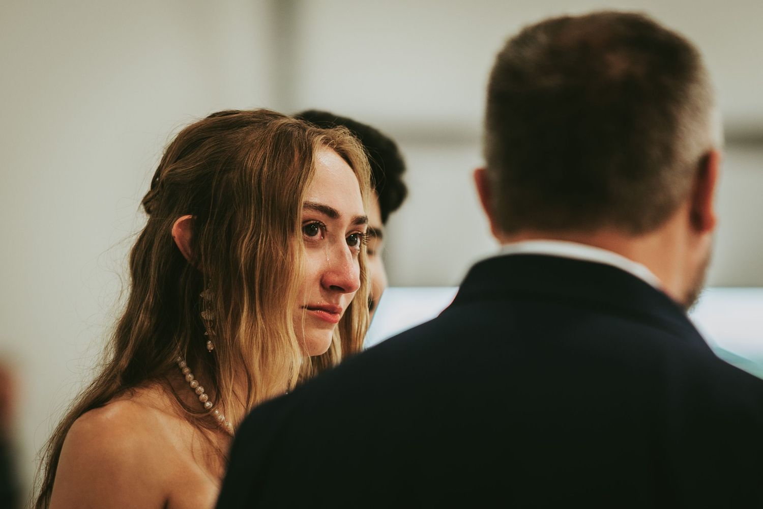 Emotional bride with tears in her eyes during wedding ceremony, wearing pearl necklace and drop earrings.