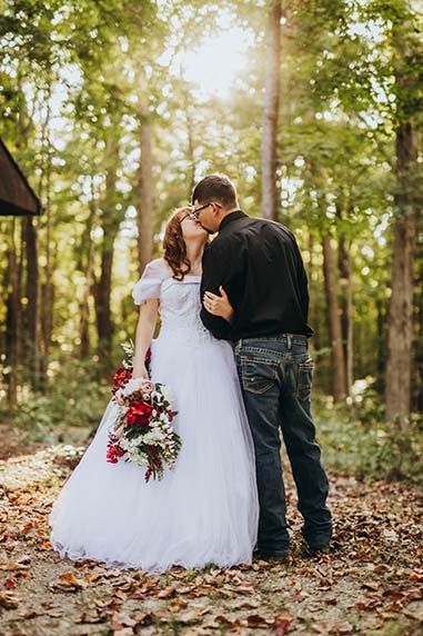 A couple in wedding attire embraces on fallen autumn leaves in a sunlit forest setting.