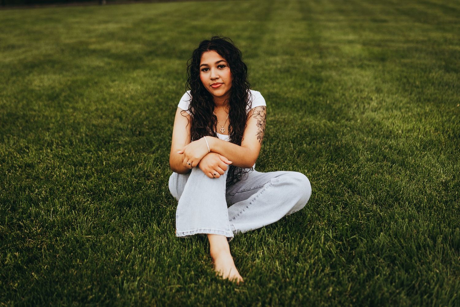 A person in white clothing sits gracefully on lush green grass in a sunlit outdoor setting.