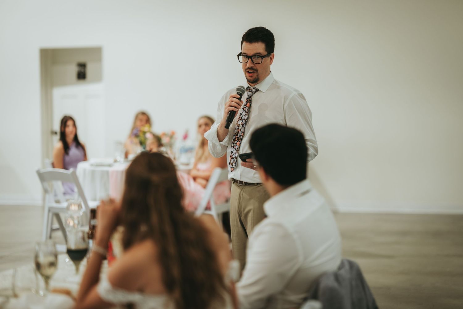 A man in glasses and a floral tie delivers a heartfelt wedding speech to seated guests in a bright reception hall.