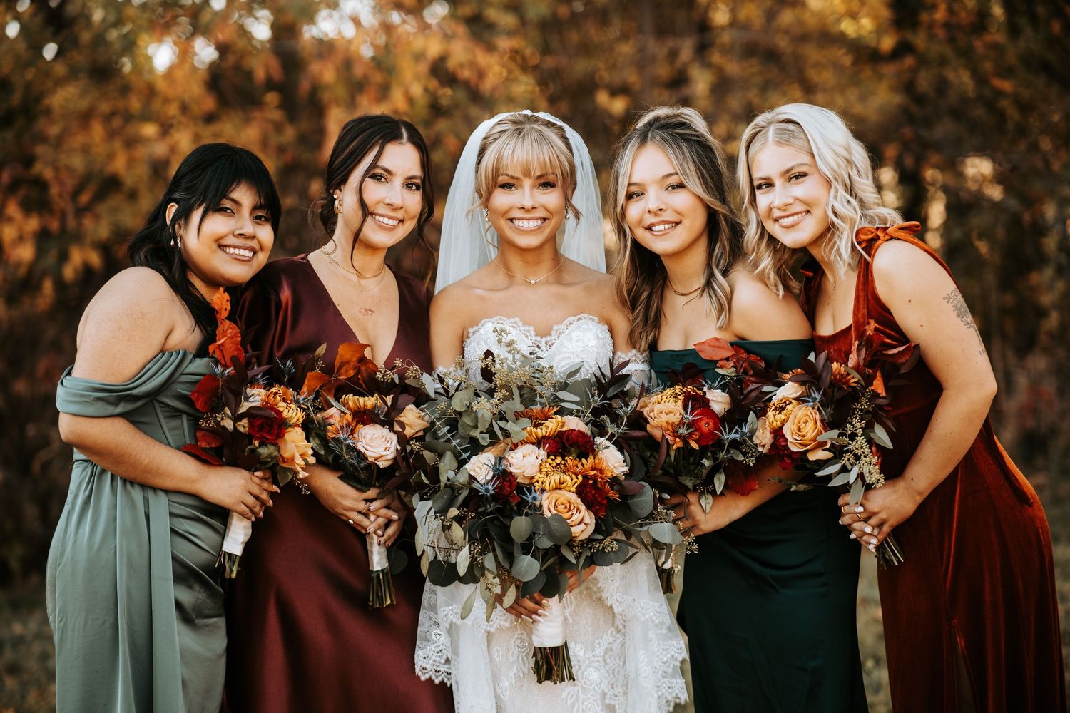 Wedding party in autumn colors holding vibrant bouquets with burgundy and orange flowers against fall foliage backdrop.