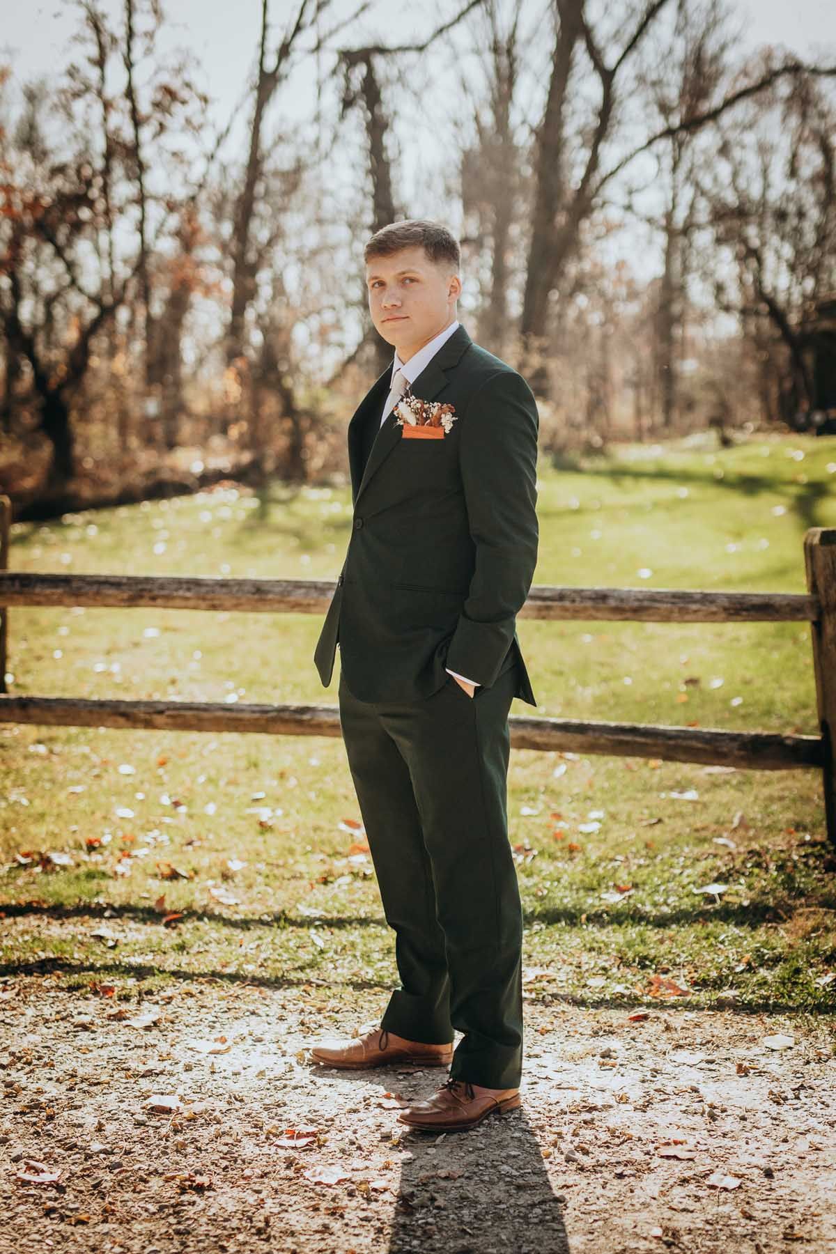 A person in a dark suit stands outdoors in autumn with fallen leaves and trees in the background.