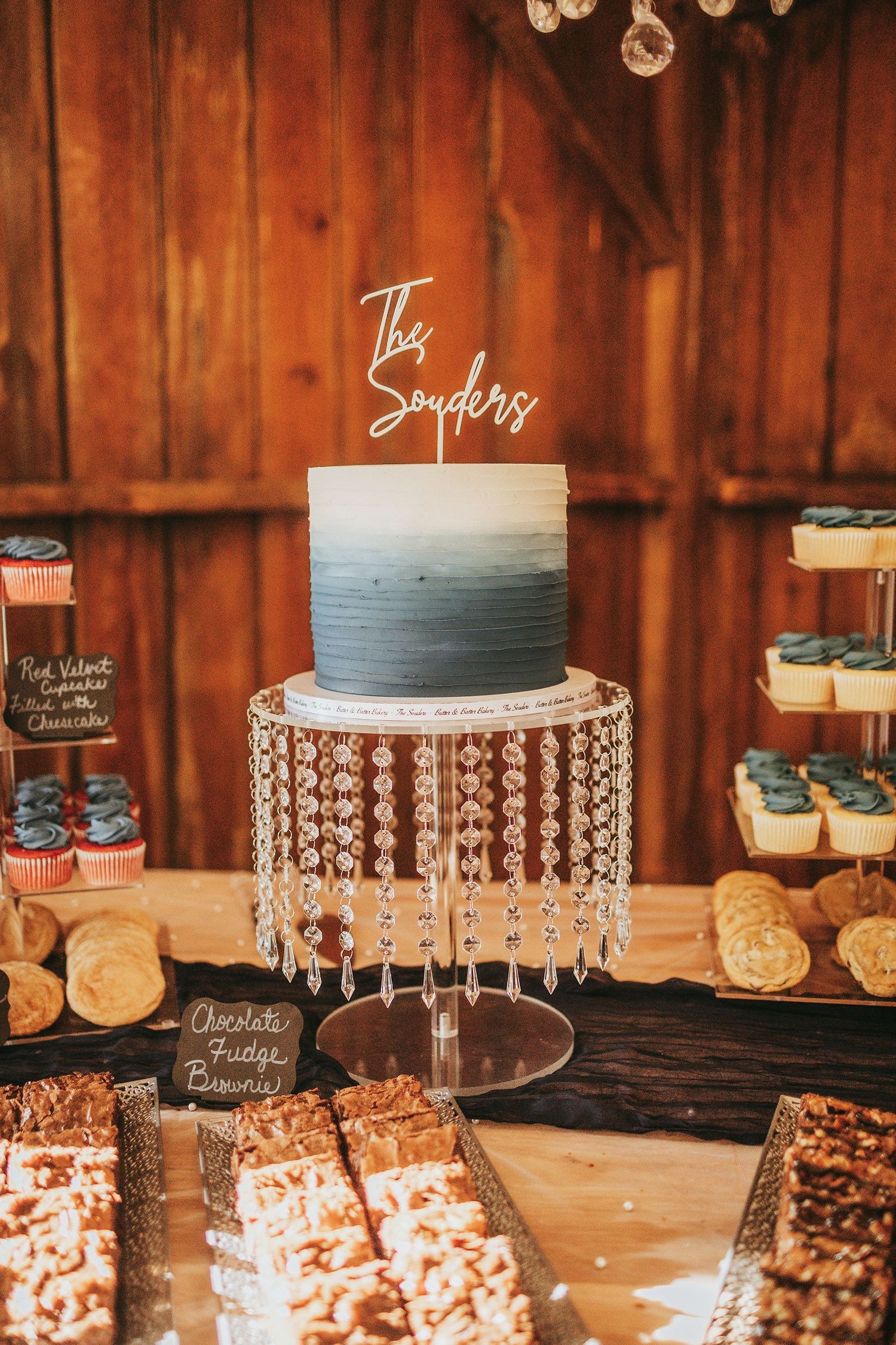 Wedding dessert display featuring elegant cakes on crystal stands with rustic wood backdrop.