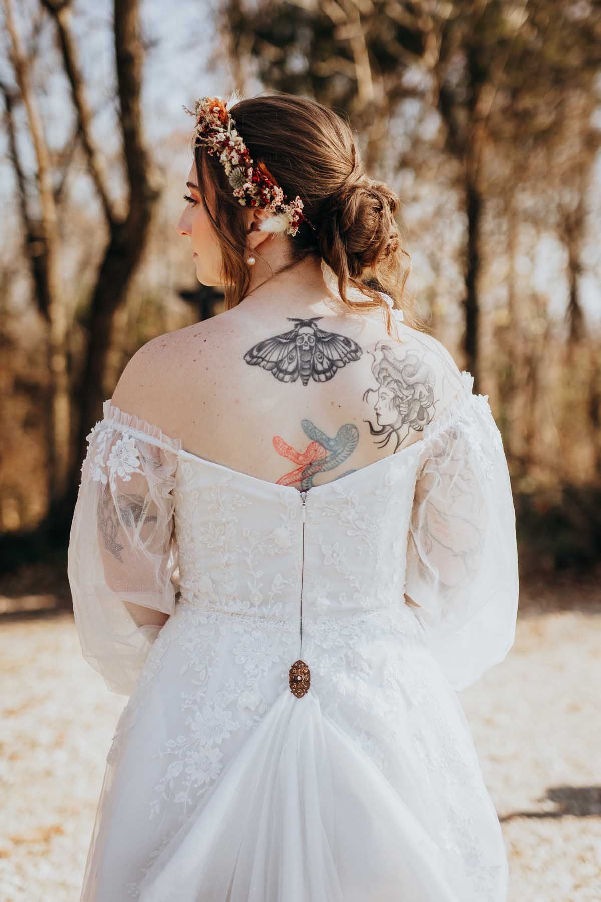Close-up view of intricate back tattoos visible through delicate white lace wedding dress details in autumn setting.