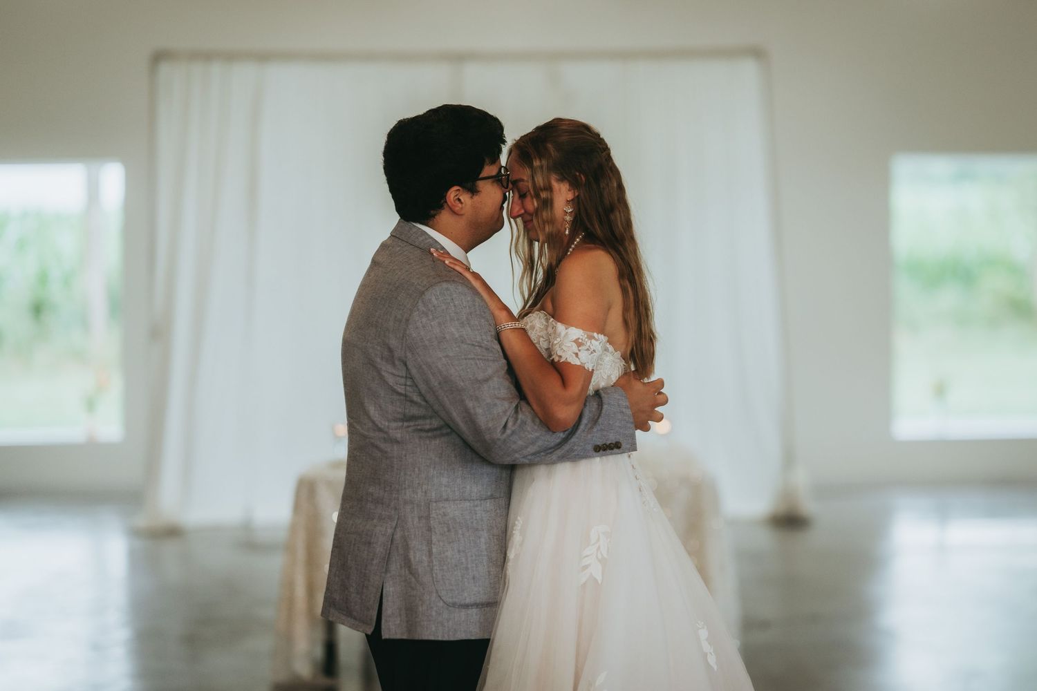 A newlywed couple shares an intimate first dance embrace in a softly lit white wedding reception venue.