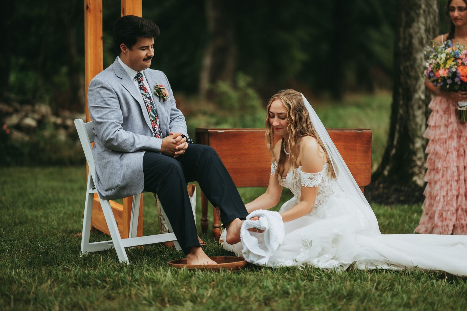 Bride and groom share a joyful moment during outdoor wedding ceremony, bride in white dress seated on grass.
