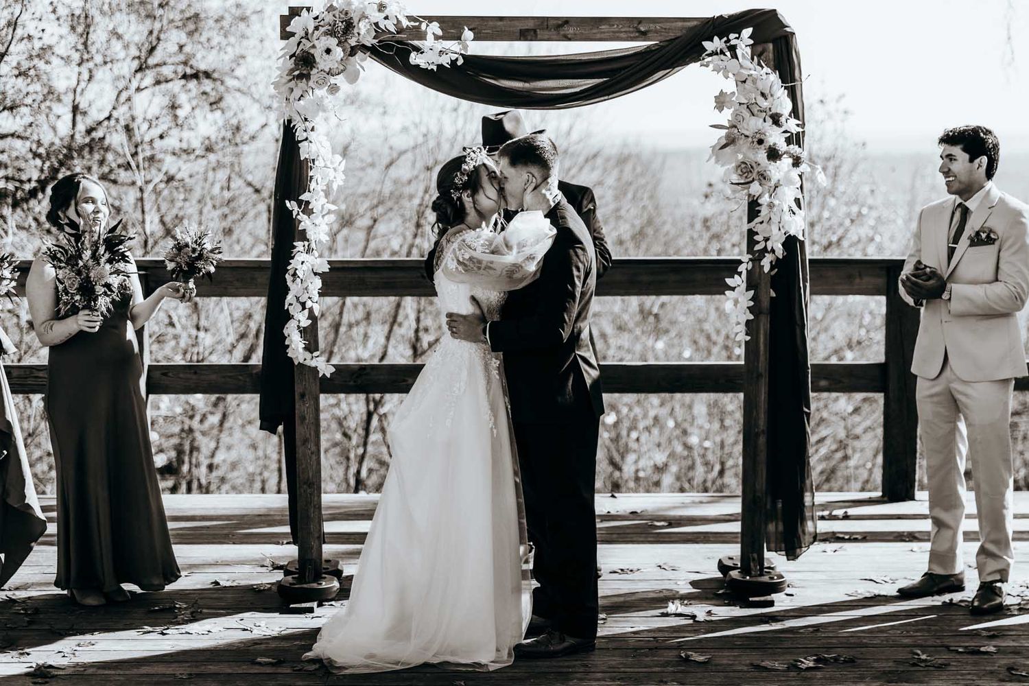 An outdoor wedding ceremony takes place under a wooden arch with guests seated on white chairs in a natural setting.