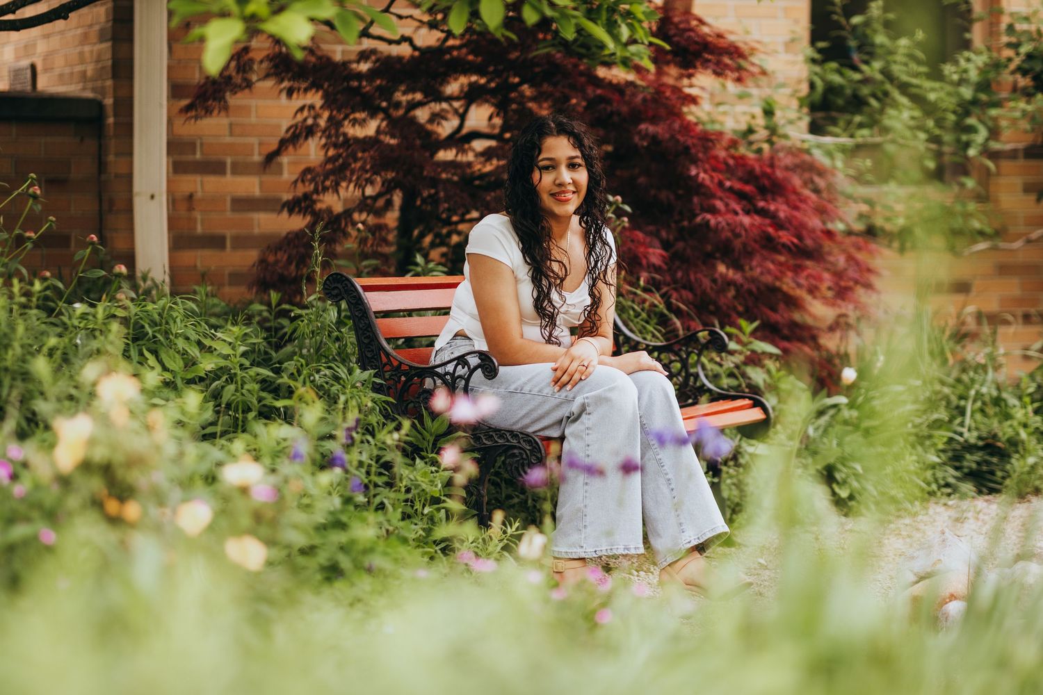 A person sits on a wooden bench in a lush garden with red Japanese maple and green foliage in the background.