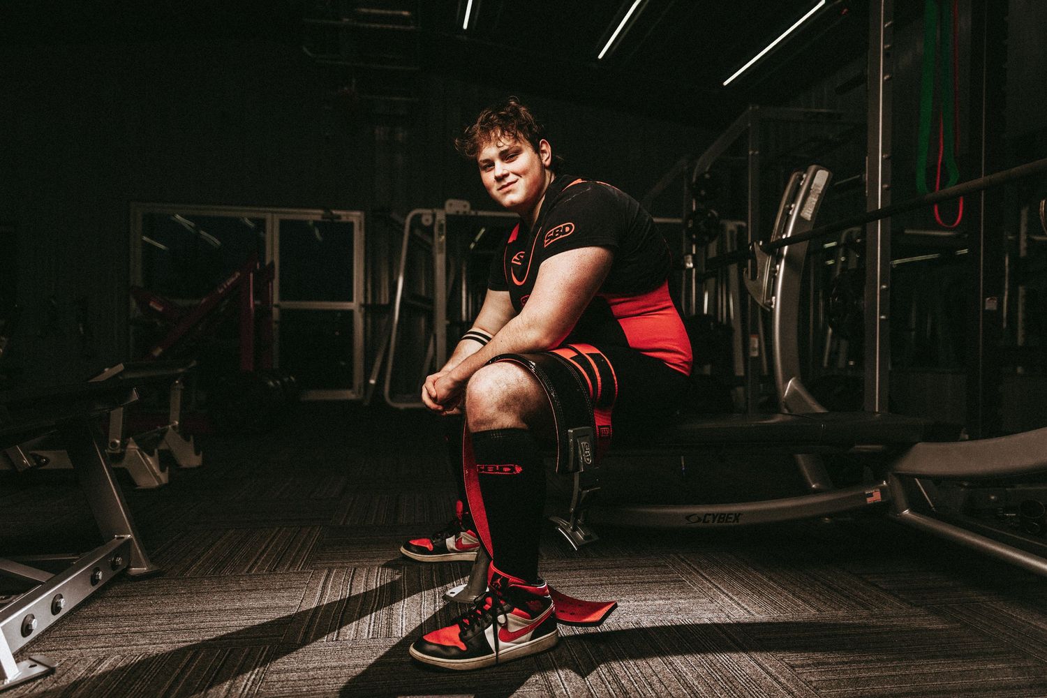Athletic person performs a dynamic stretching exercise in a dimly lit gym setting with workout equipment visible.