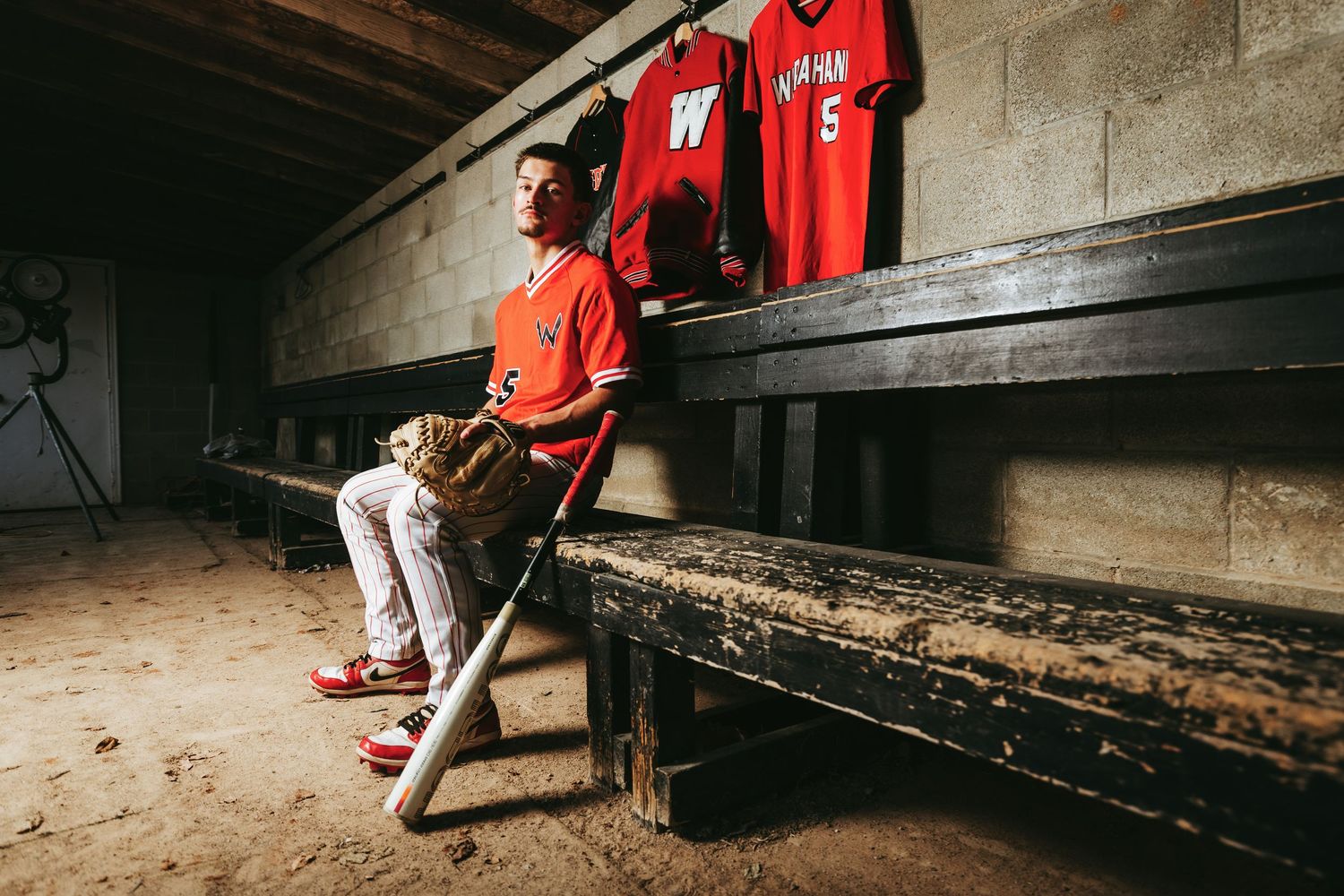 Baseball player in red uniform sits in shadowy dugout with concrete wall behind.