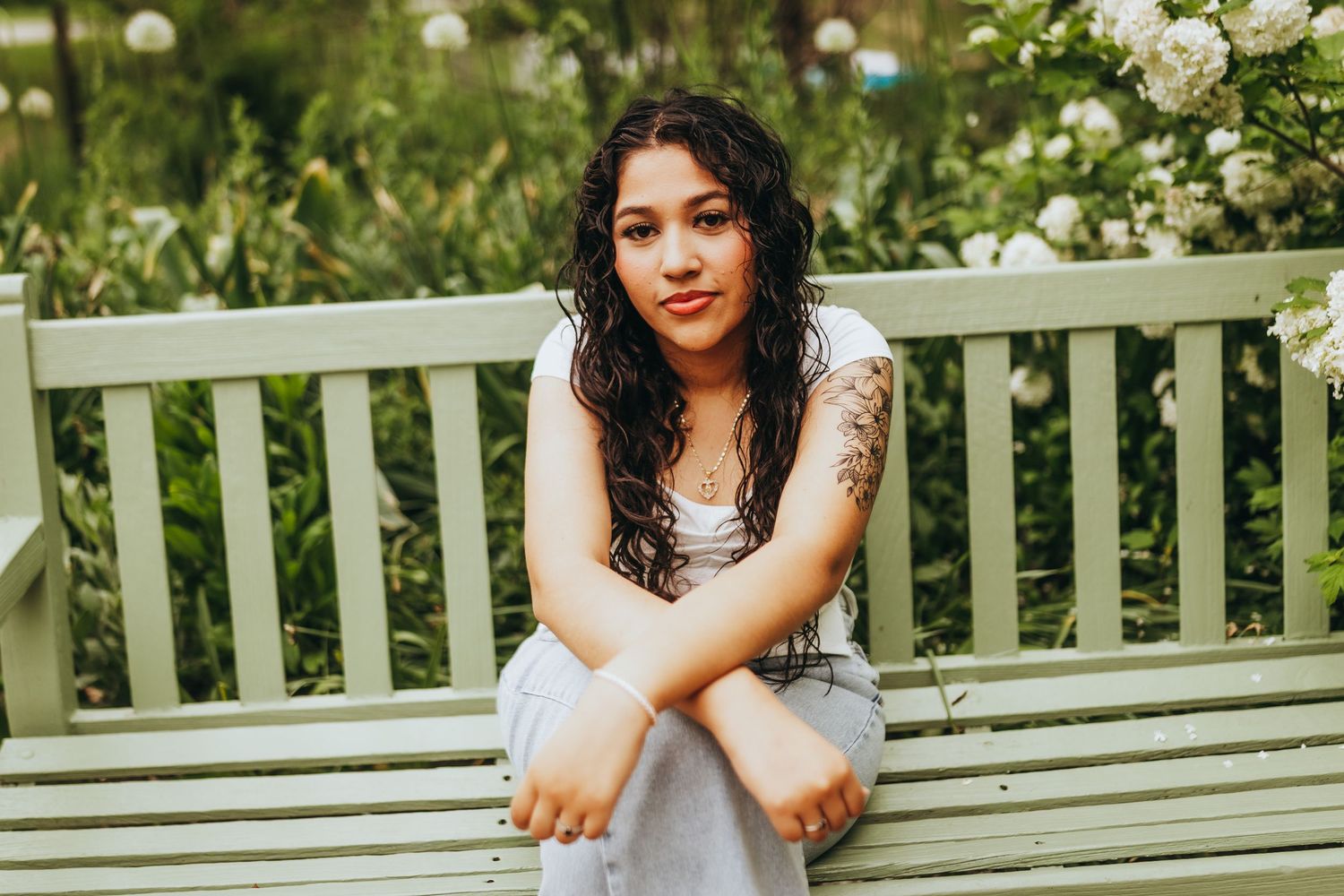A person in a white dress sits on a green garden bench surrounded by blooming white flowers and greenery.
