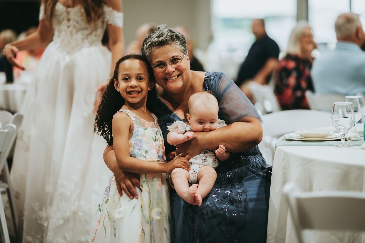 Woman smiling with young flower girl and baby at wedding reception.