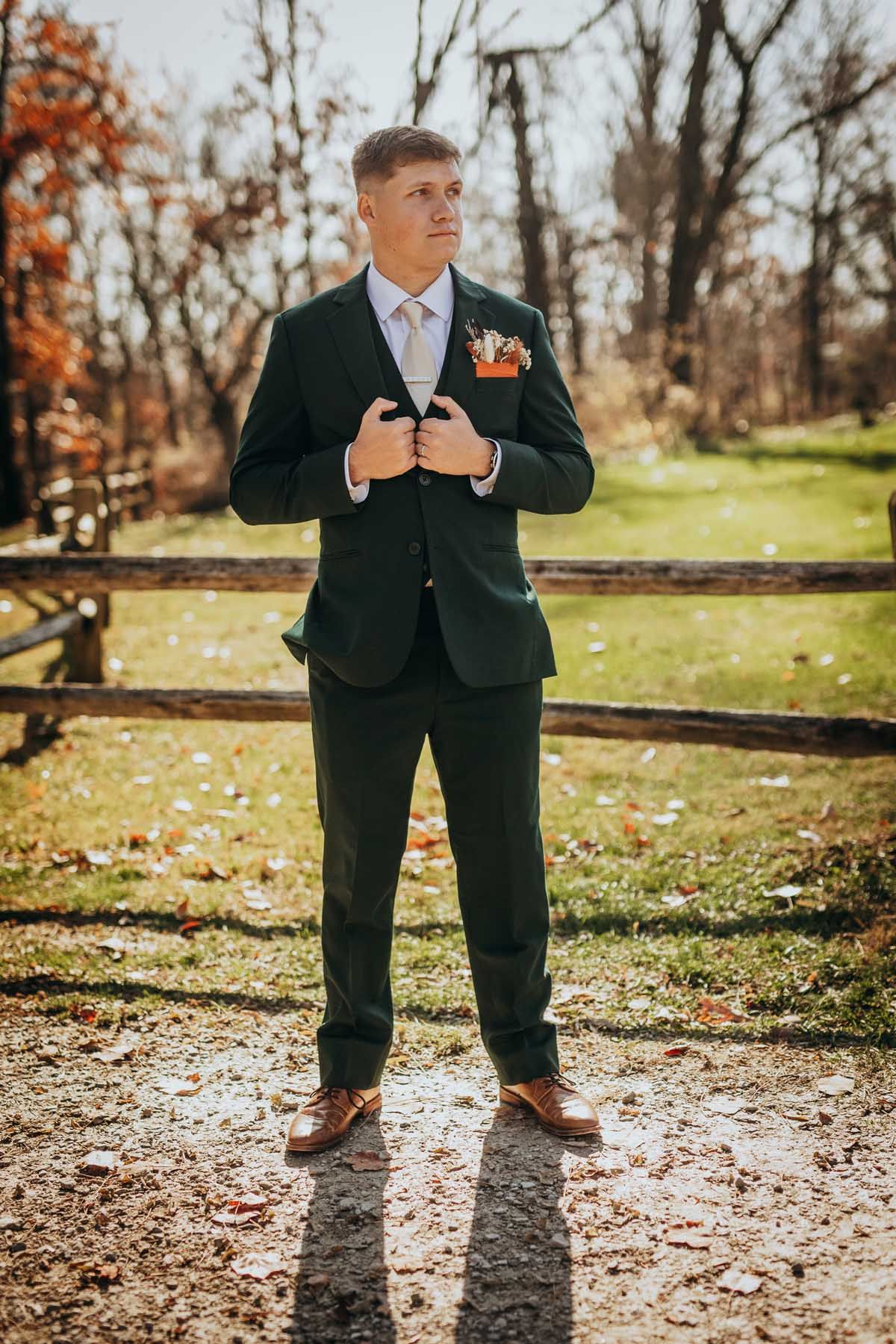 A person in a black suit and brown shoes stands on fallen autumn leaves on a sunny day in a park setting.