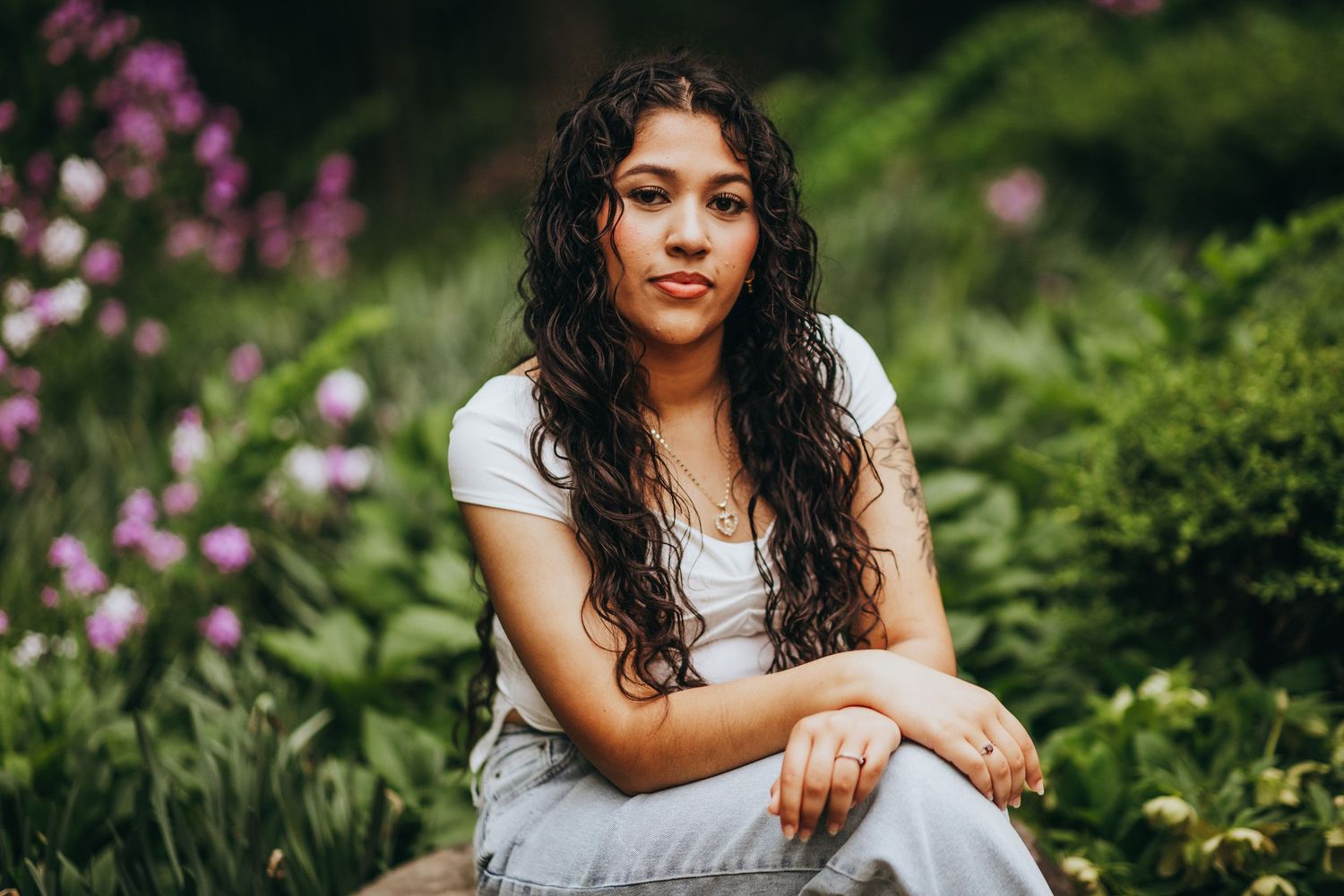 A person with long dark curly hair sits in a garden with blooming pink flowers and lush greenery in the background.