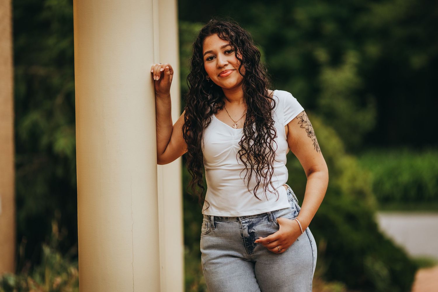 Woman in gray top and jeans poses outdoors in natural lighting against blurred green background.