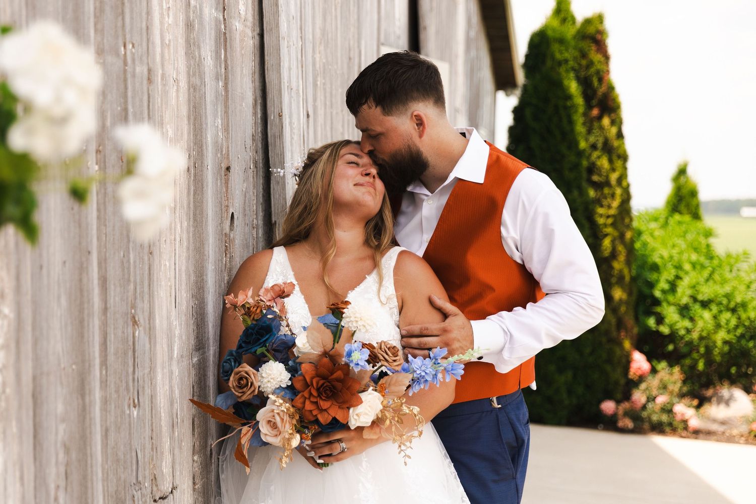 A couple in wedding attire share a romantic moment against a rustic wooden fence backdrop in a garden setting.