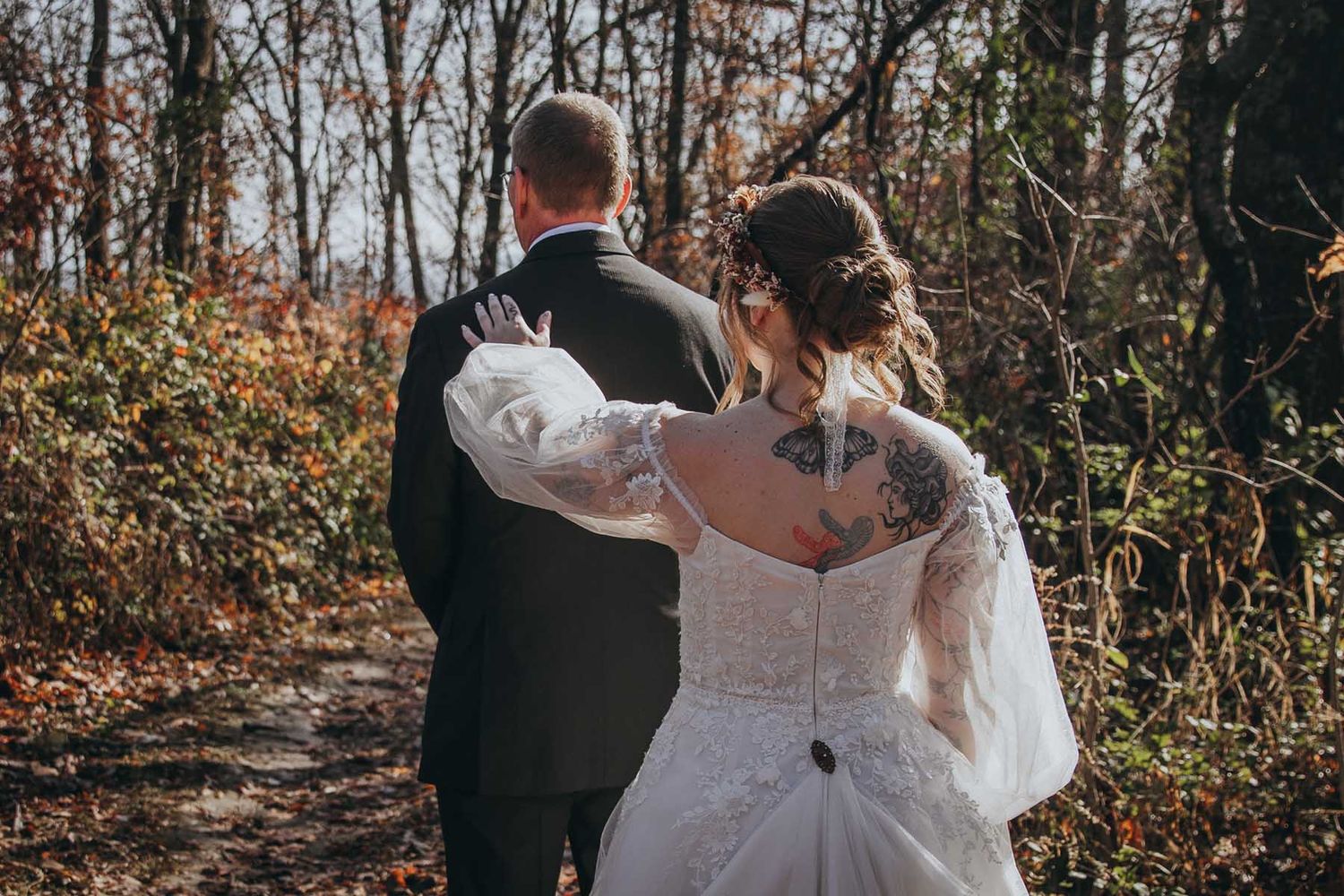 Couple in wedding attire embraces while walking through a sunlit forest path during fall season.