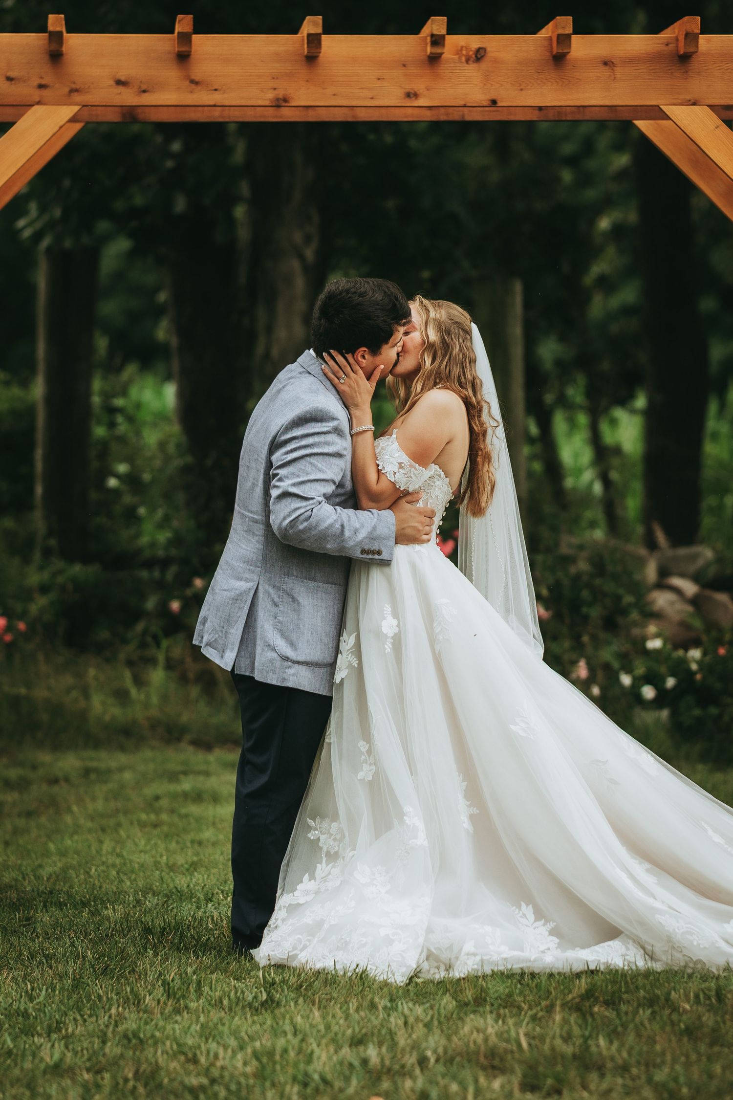 Bride and groom share their first kiss under a wooden pergola arch surrounded by lush greenery at an outdoor wedding.