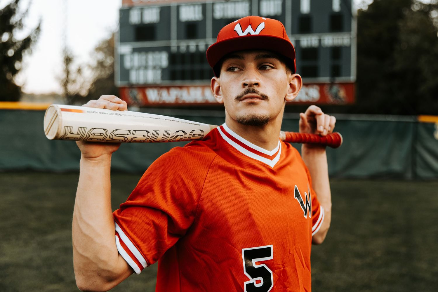 Baseball player in orange uniform and black cap poses with bat against blurred outdoor background.