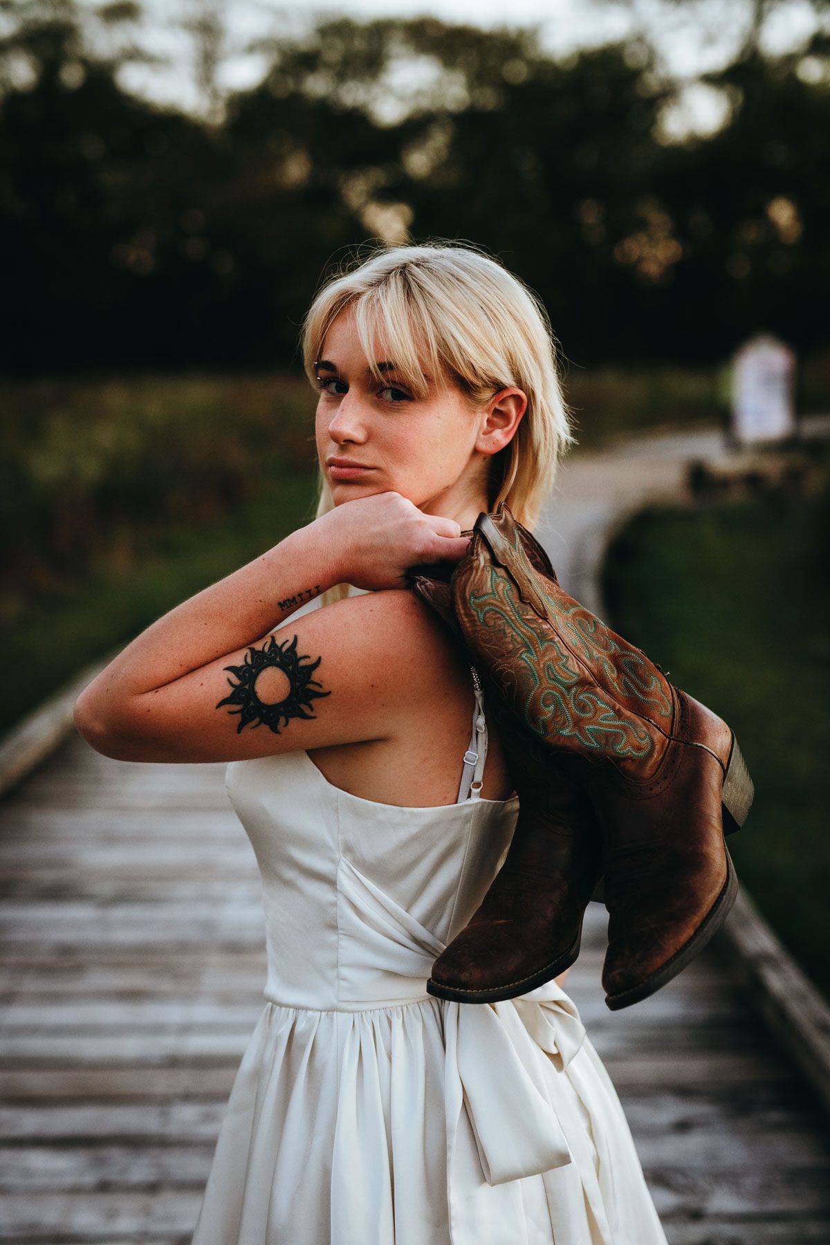 Woman in white dress with sun tattoo and brown jacket poses on wooden boardwalk at dusk.