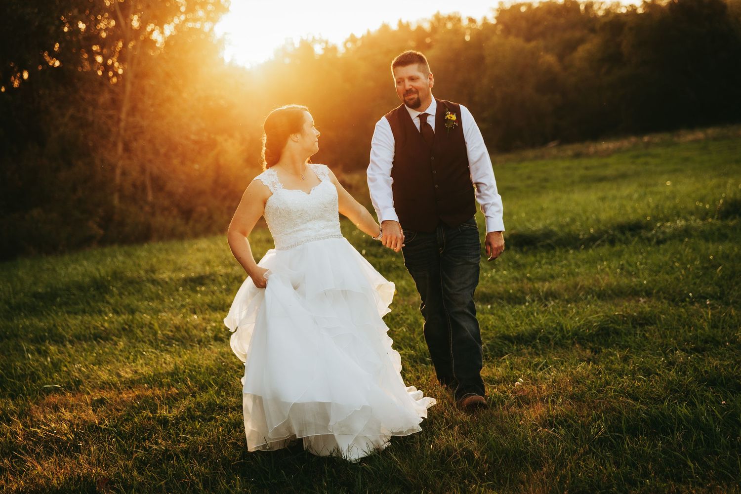 A couple in wedding attire walks through a golden sunset field with warm glowing light illuminating their silhouettes.
