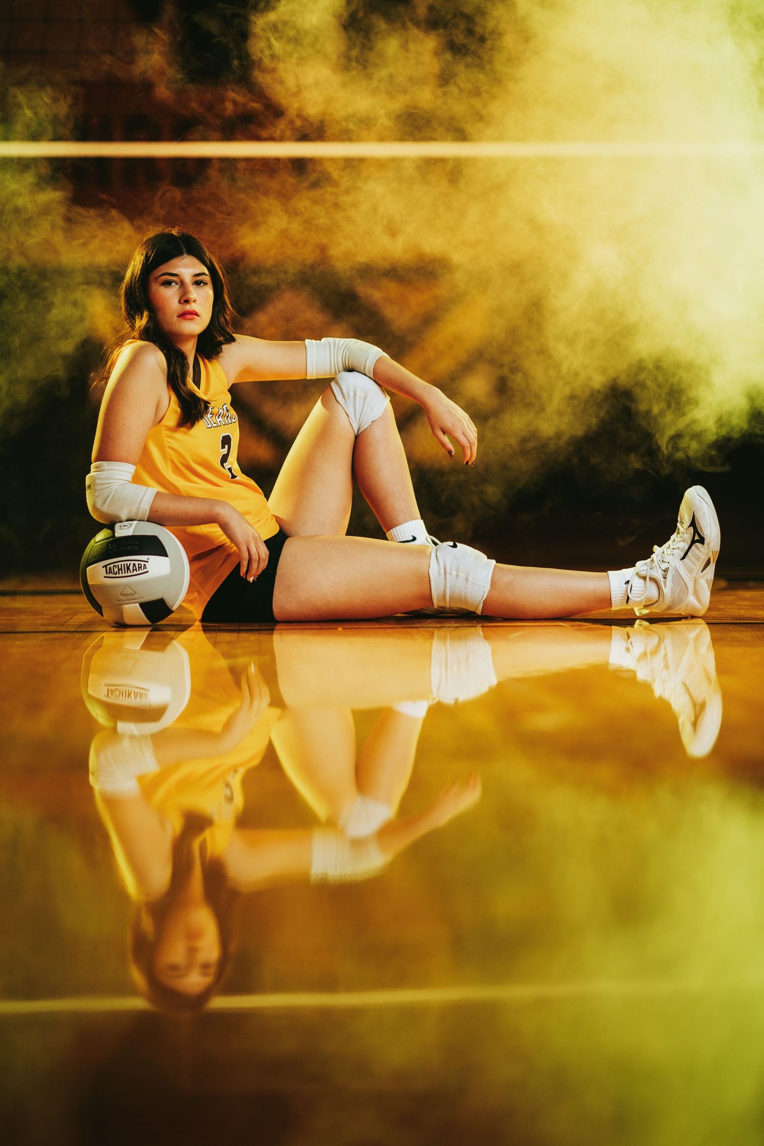A volleyball player in yellow uniform sits on reflective court floor with dramatic yellow smoke effect in background.