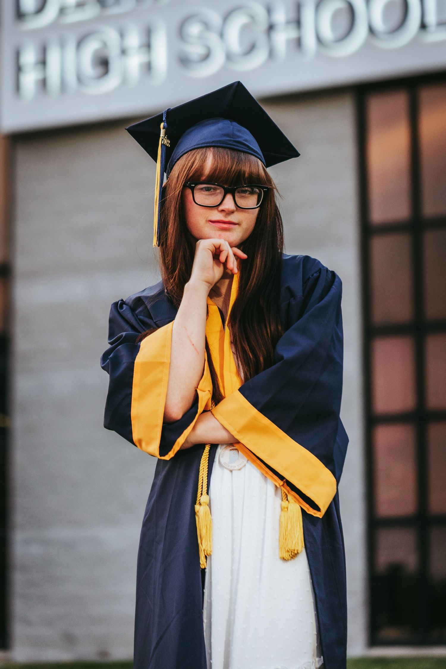 A graduate in navy cap and gown with yellow stole poses for a series of photos in front of a school building.