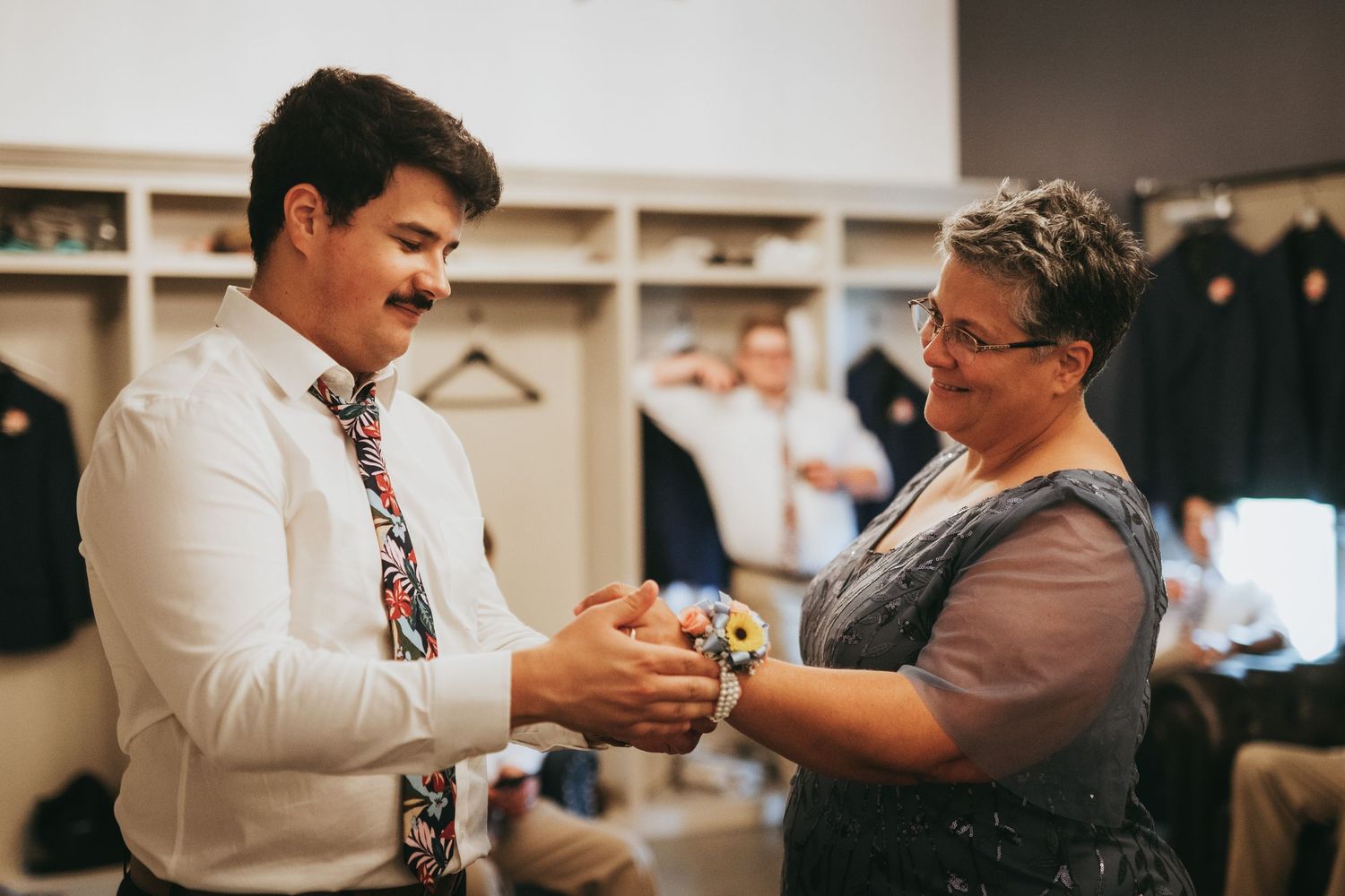 A young man in a white shirt and floral tie shares a tender moment with an older woman wearing a corsage at a wedding.