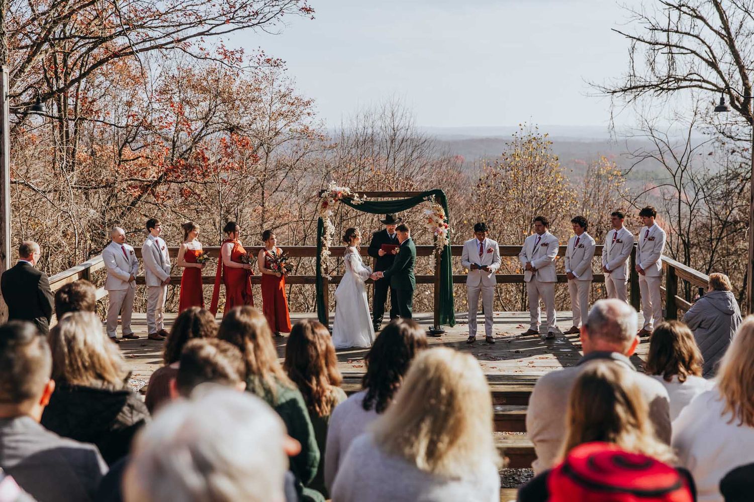 A crowd gathers outdoors for a wedding ceremony on a scenic overlook during autumn with bare trees in the background.