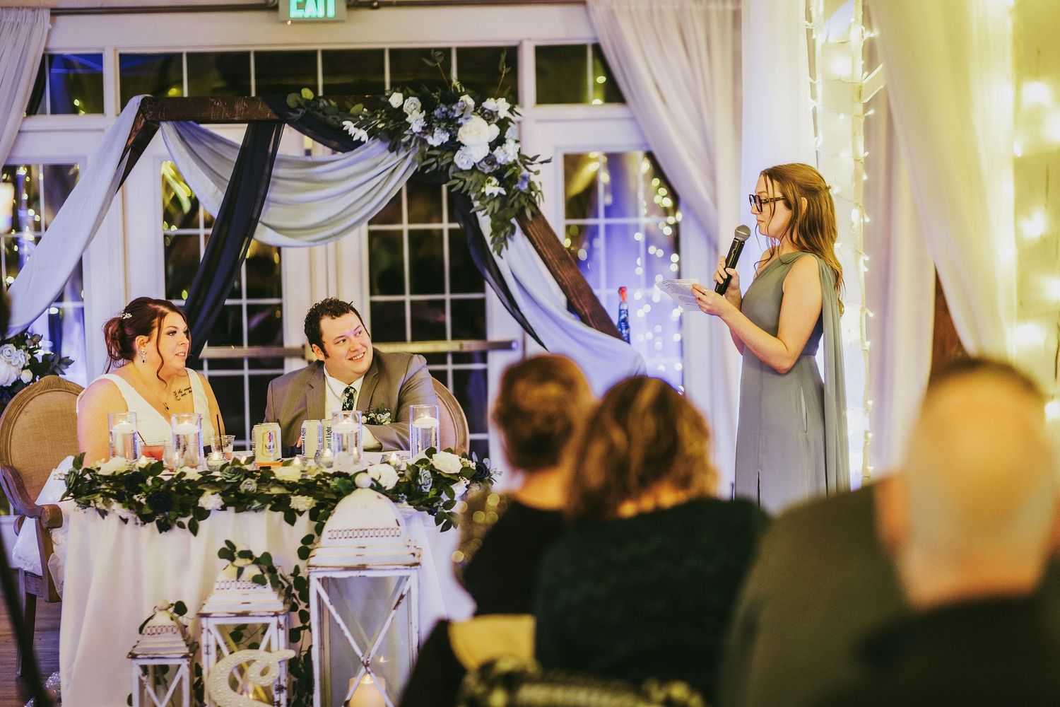 A wedding reception speech moment in a venue decorated with white draping and floral arrangements.