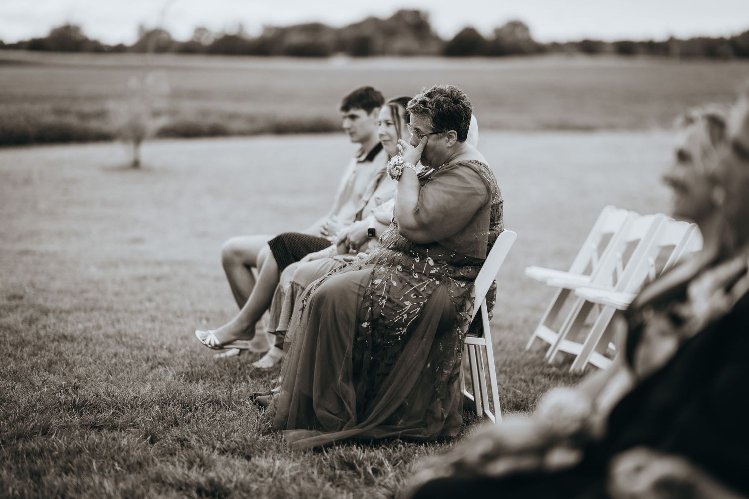 Wedding guests seated on white chairs in outdoor ceremony on grassy field, black and white photo.