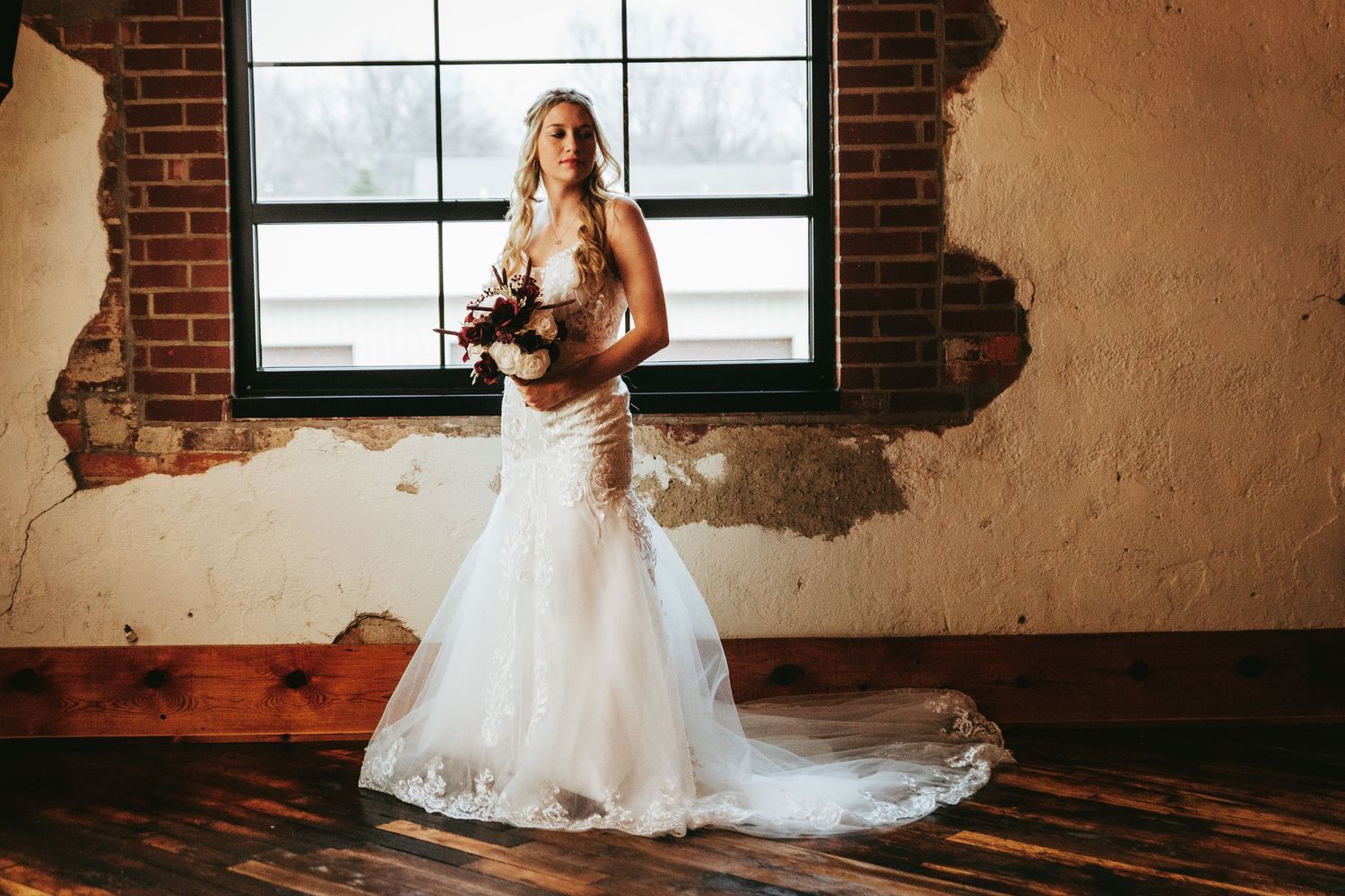A bride in a flowing white wedding gown stands elegantly in front of rustic brick walls and windows.