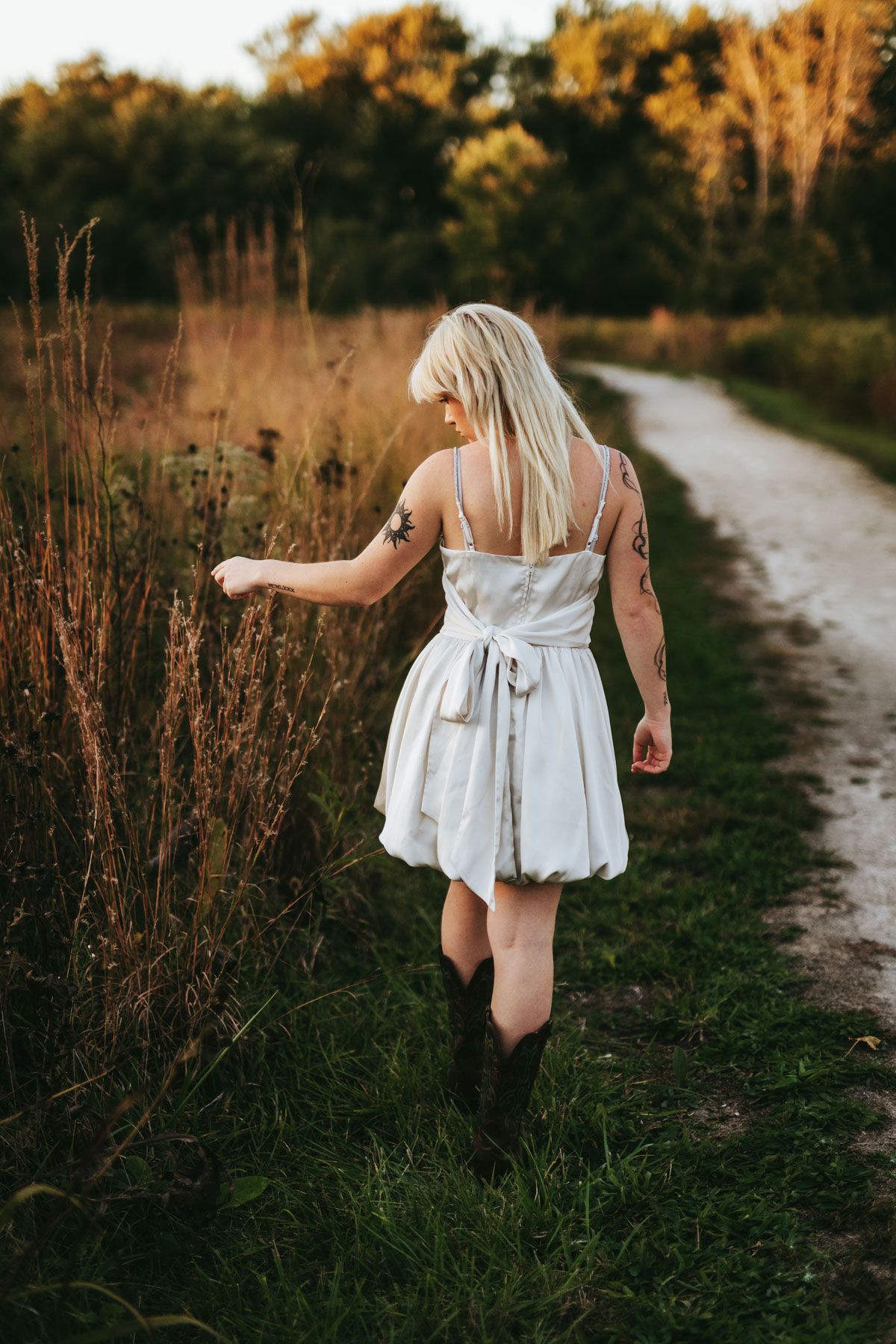A person in a flowing white dress walks along a winding dirt path through tall grass at golden hour.