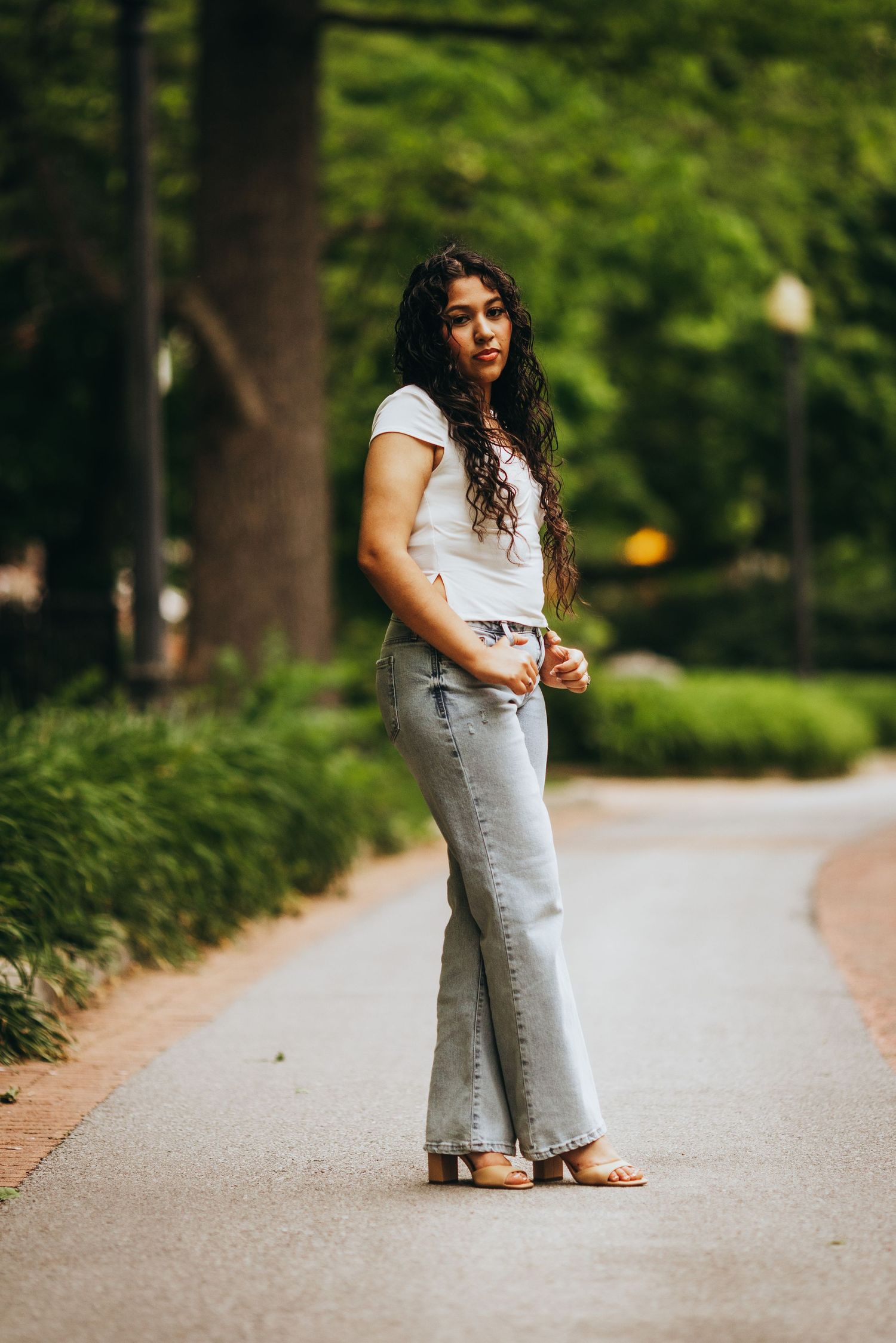 A person in a white top and light denim skirt walks along a tree-lined path in a park on a sunny day.