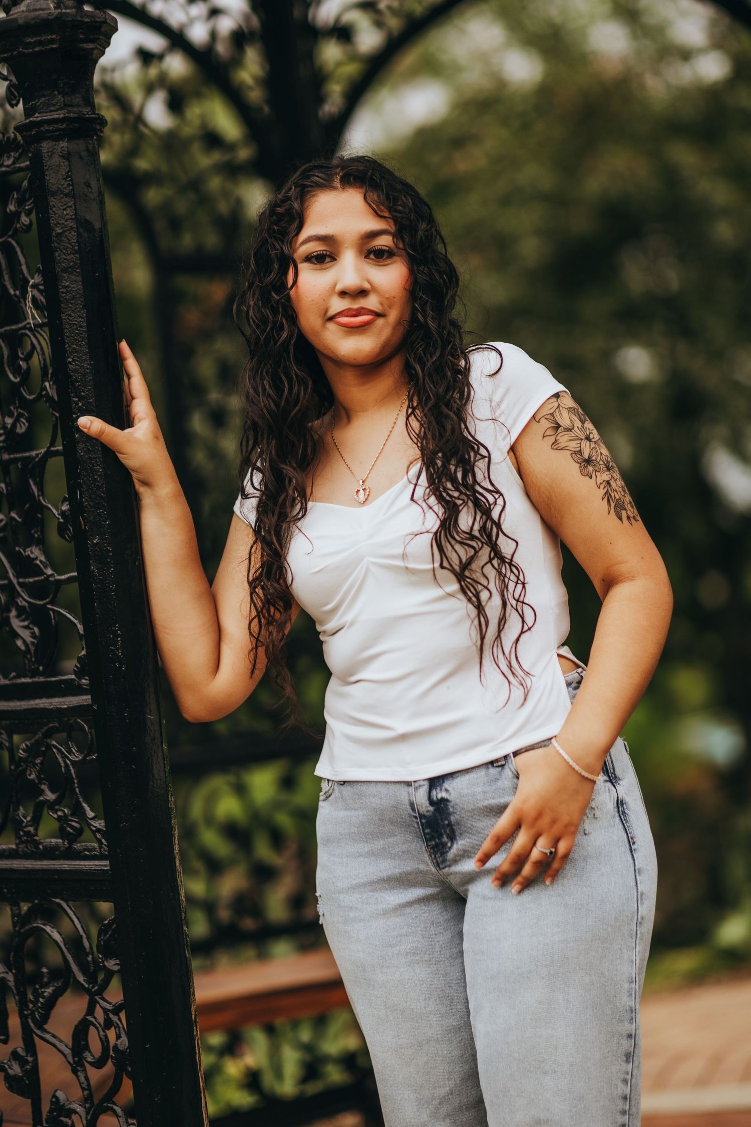 Casual outdoor portrait with white t-shirt, light jeans and visible tattoos leaning against ornate iron railing.