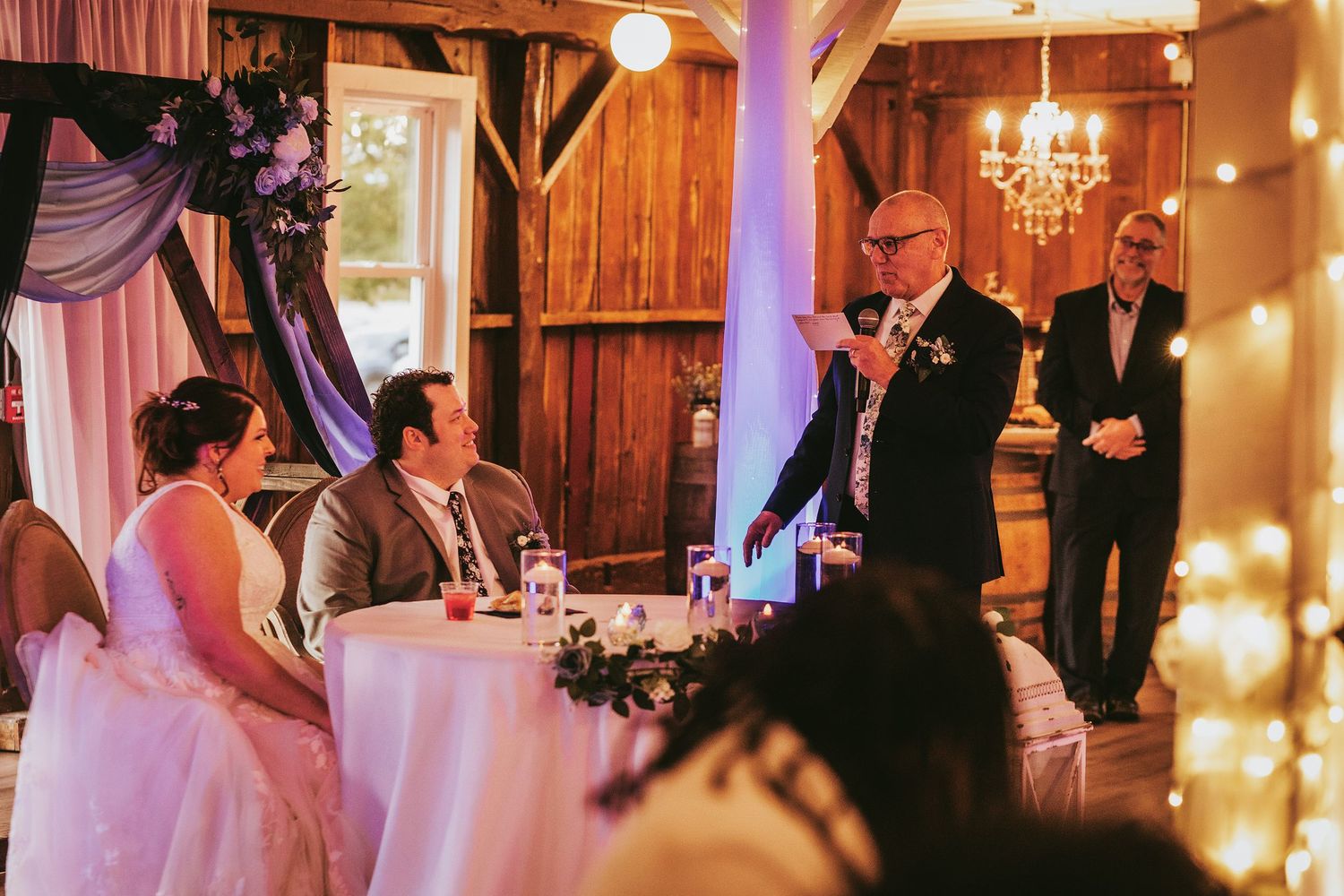 A wedding toast being given in an elegant rustic venue with wooden walls and a crystal chandelier.
