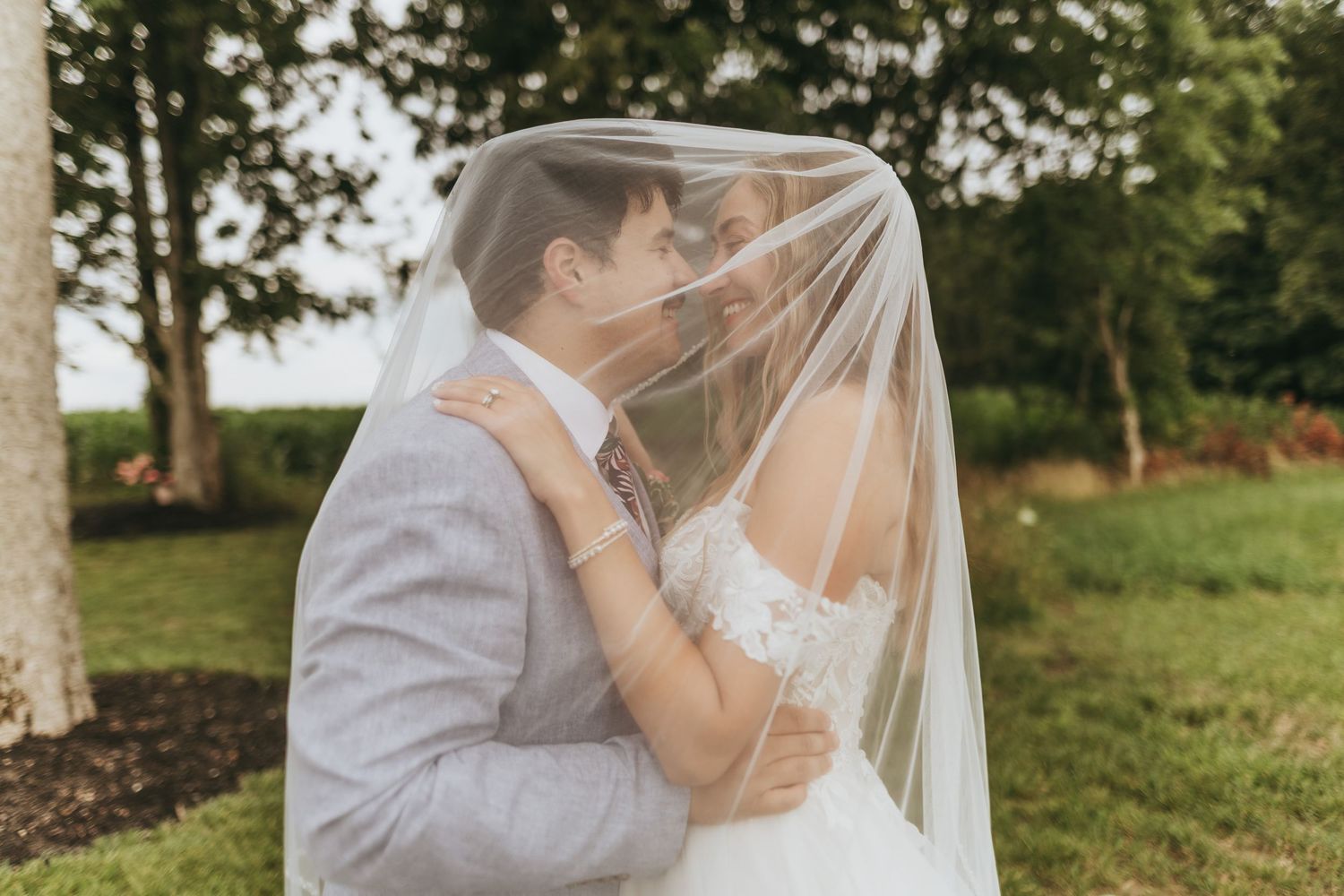 Bride and groom sharing romantic kiss under flowing white veil in lush garden setting.